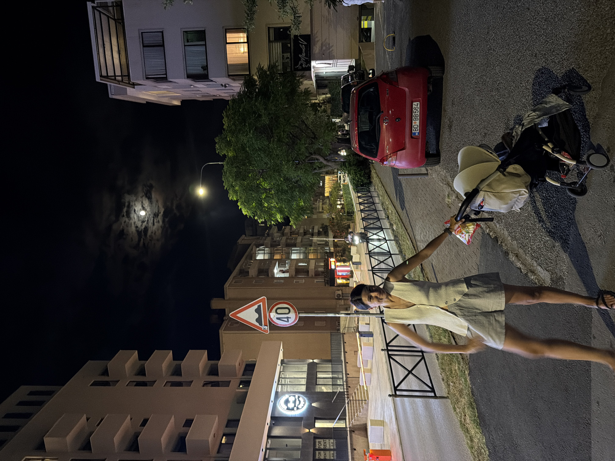 The image shows a night scene in what appears to be a residential or commercial area. In the foreground, a young woman is captured in motion, her arms outstretched and holding a bag of snacks. She is wearing a light-colored sleeveless top and shorts, with sandals on her feet. Her expression is joyful and energetic. Behind her, a stroller is parked on the asphalt, with a baby carriage component visible. To the left, a red car is parked, with a license plate that reads "BD BB 564". Further in the background, buildings of various heights are illuminated, suggesting it's nighttime. A large tree stands prominently near the red car, its leaves illuminated by nearby lights. The sky features a prominent full moon surrounded by clouds, indicating a clear night. Traffic signs are visible, including a sign with "40" indicating a speed limit. A lamppost with an illuminated bulb is also present. The scene is busy with architectural elements and signs of urban life.
