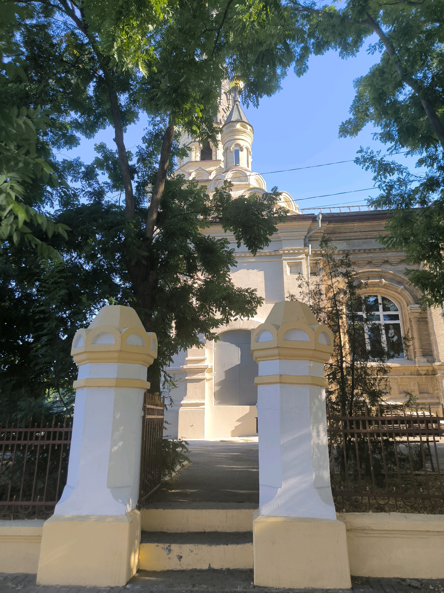 The image shows the entrance to the Church of St. Nicholas the Wonderworker in Mykolaiv, Ukraine. The church, a prominent landmark in the city, is partially obscured by lush green trees. In the foreground, two ornate white and yellow pillars flank a set of steps leading to the church. A dark metal fence runs along the sides of the pathway. The church itself is a light yellow color with classical architectural details, including arched windows and decorative moldings. The sky is a clear, bright blue, suggesting a sunny day. The overall impression is one of peace and solemnity.