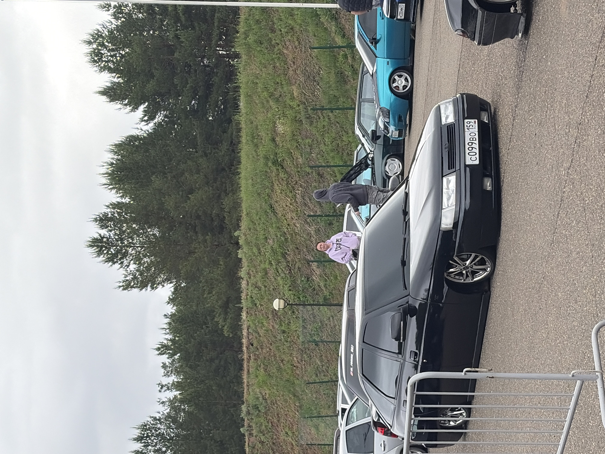 The image captures an outdoor scene in Прикамский, Russia, featuring a gathering of multiple modified automobiles on a paved surface. In the foreground, grey metal pedestrian barriers partially obscure a black two-door sedan, heavily lowered with chrome multi-spoke wheels, displaying a Russian license plate "C099BO 159". To its left, a silver vehicle is partially visible. Behind the black sedan, a dark green sedan with its hood open and white multi-spoke wheels is parked next to a turquoise sedan, also lowered with white five-spoke wheels and a dark hood, featuring a license plate "M330MM 73".
Several individuals are present among the cars. A person in a light purple hooded sweatshirt stands in the mid-ground on the left, looking towards the right. Another person in a dark grey hooded sweatshirt and camouflage pants stands facing away, observing the cars. A partial view of a third person wearing a dark vest is visible on the far right.
The background features a chain-link fence separating the paved area from a rising grassy slope. A single street lamp with a spherical white globe is situated on the slope. Beyond the slope, a dense line of coniferous trees forms the horizon under an overcast, light grey sky. A thin white vertical pole is visible along the far right edge of the image.