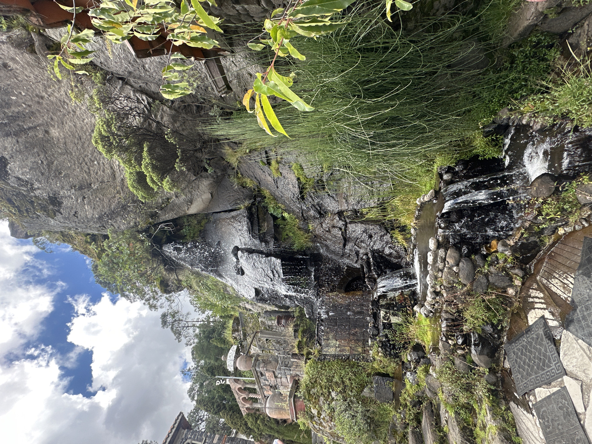 The image depicts a multi-tiered waterfall cascading down a rocky, moss-covered cliff face. The water flows into pools and streams that are bordered by stones and lush green vegetation, including tall, reedy grass and leafy plants. To the left of the waterfall, a stone staircase with mossy steps ascends the hillside, leading to a complex of stone structures with arched openings and large ceramic pots. The architecture appears rustic and possibly historical. In the foreground, there are several dark, patterned mats laid out on a stone patio, suggesting an outdoor seating or resting area. The sky above is partly cloudy with bright white clouds against a blue background. The scene is set in a natural, yet cultivated, environment, potentially a garden or a historical site in San Mateo Ozolco, Mexico.
