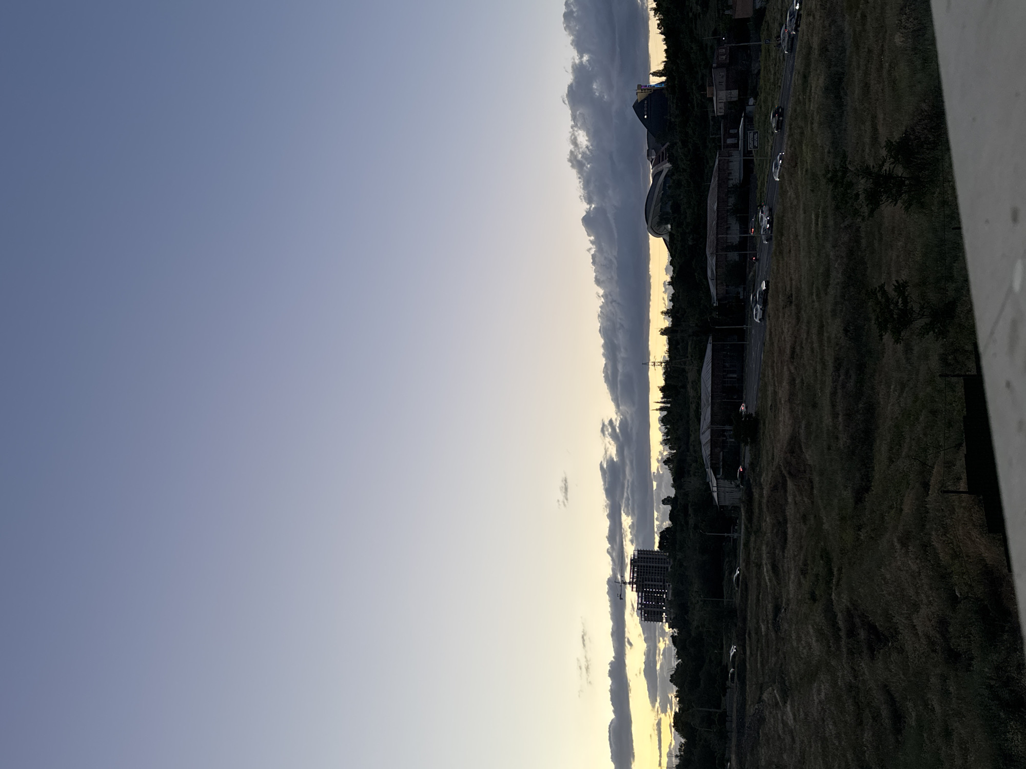 The image presents a panoramic view of an urban or suburban landscape at dusk, likely taken from an elevated position. The upper portion of the frame is dominated by a vast sky that gradients from a deep periwinkle blue at the top-left to a pale yellow and light gray closer to the horizon, indicative of twilight. A prominent, elongated band of dark, dense clouds stretches horizontally across the middle of the sky, just above the distant landscape.
Along the distant horizon, a dense line of dark trees is visible. Several buildings punctuate this skyline: on the far left, a distinctive, modern-style building with a curved roof and illuminated sections stands out. Further to the right, a multi-story building under construction with visible scaffolding is present. Below the horizon, a road can be seen with several vehicles, some showing faint reflections from their lights as they travel. The area between the road and the distant horizon is covered in patchy, dry-looking vegetation.
The immediate foreground, particularly on the right side, consists of extensive areas of dry, unkempt grass and scattered dark trees. A light-colored concrete barrier or wall is partially visible on the far right edge. No people are discernible, and no visible text can be read in the image. The scene captures the quiet, transitional light of early evening in what is identified as Yerevan, Armenia, with elements of both developed areas and natural, untended land.