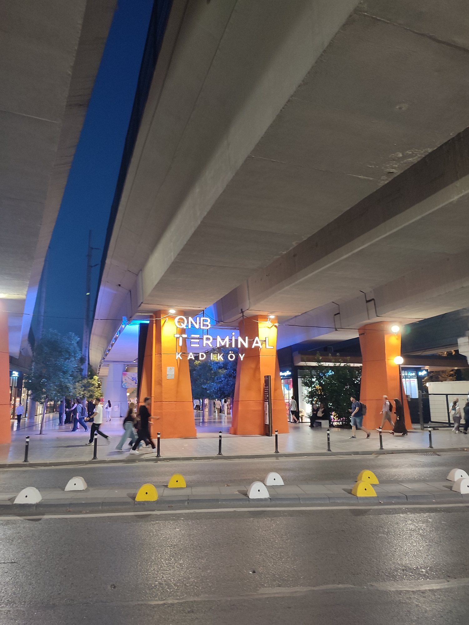 The image shows a view of the QNB Terminal Kadıköy in Istanbul, Turkey, at night. The terminal is characterized by its bright orange pillars and illuminated signage. Above the terminal, a large concrete overpass casts shadows, with a section of deep blue sky visible between the concrete structures.
Several people are walking on the sidewalk in front of the terminal. Some are walking individually, while others are in small groups. The street in the foreground has a row of white and yellow concrete traffic barriers. The lighting suggests it is dusk or early evening.
