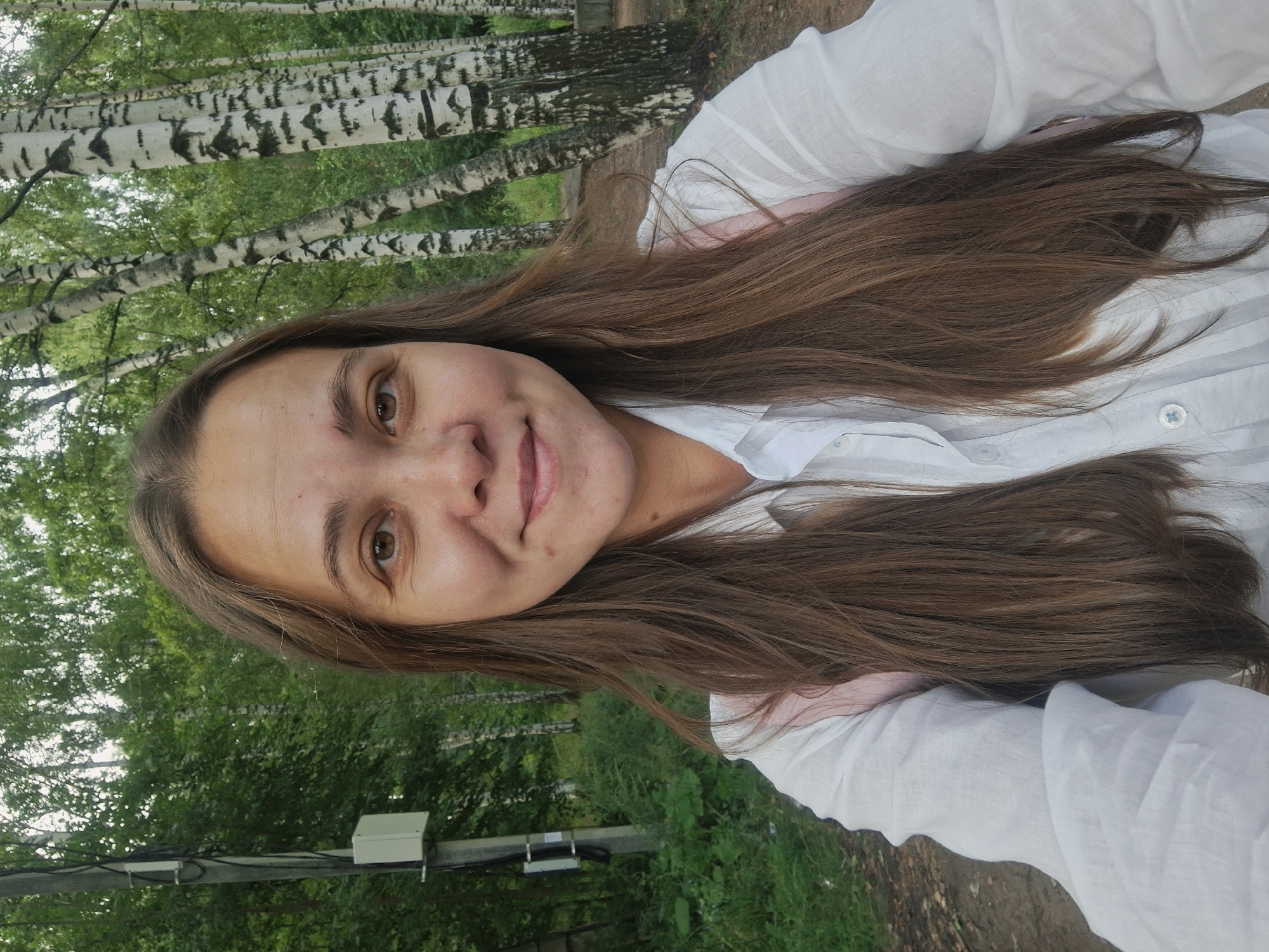 A young woman with long, brown hair smiles at the camera in an outdoor setting. She is wearing a white, collared shirt. Behind her, a line of birch trees with white bark and black markings is visible, along with lush green foliage. A utility pole with a small white box attached is situated to the left of the woman. The ground appears to be a dirt path with some scattered debris. The overall scene suggests a park or a wooded area within a city. The location is identified as Chaikovsky, Russia.