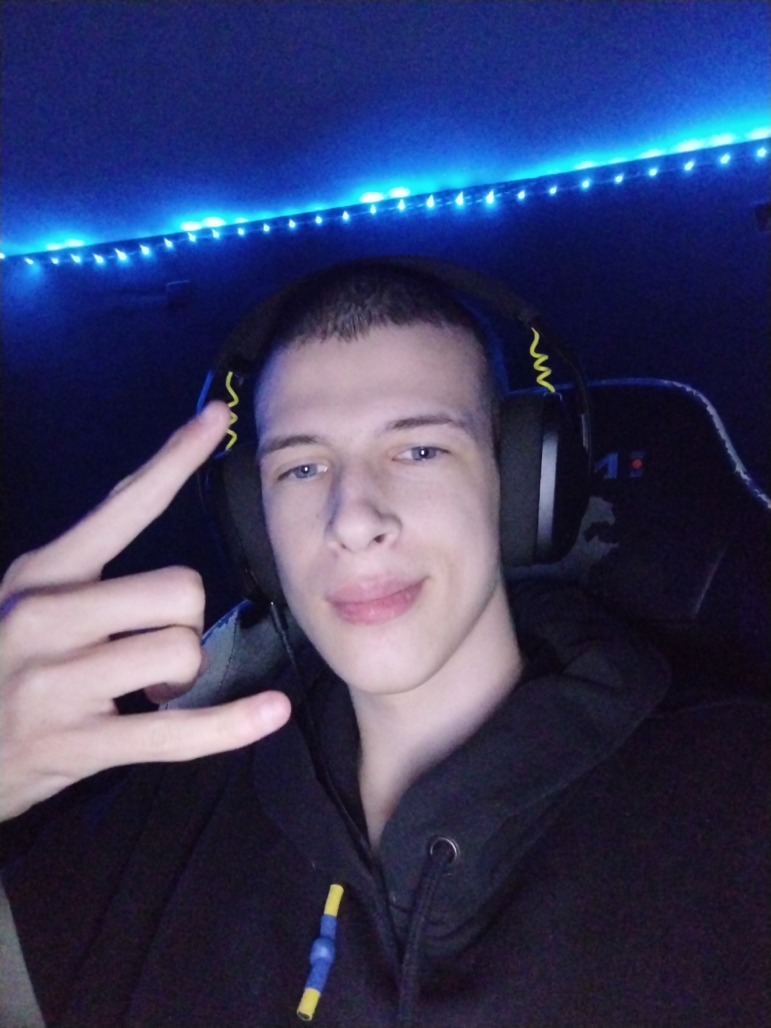 The image features a young man, likely in his late teens or early twenties, depicted indoors in a low-light setting. He has short hair, blue eyes, and a slight, relaxed smile. He is wearing a black hoodie and black over-ear headphones, which are notable for a distinct yellow coiled cable design on the earcups.
His left hand is raised, forming a "rock on" or "devil horns" gesture with his index and pinky fingers extended and his thumb also visible. On the front of his hoodie, a small yellow and blue object, possibly a toggle or decorative pull, is visible. Behind him, the background is dark, primarily illuminated by blue LED strip lights running horizontally along the top edge of a wall or ceiling. A dark gaming or office chair is partially visible behind his right shoulder, featuring an unreadable logo or design on its backrest.
The scene suggests a personal space, such as a bedroom or gaming room, during the evening or nighttime, indicated by the artificial blue lighting. The overall mood is relaxed. This media file originates from Svetovrachene, Bulgaria.