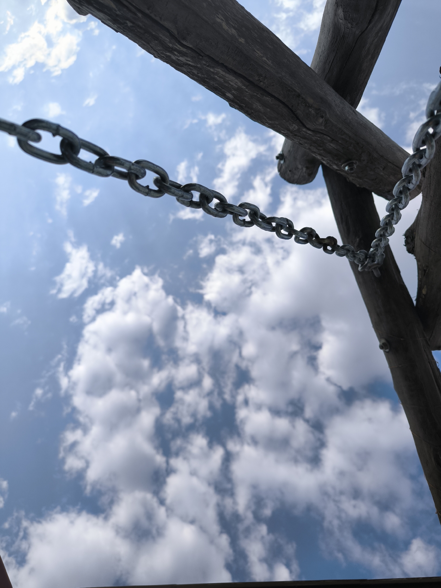 A low-angle shot captures a metal chain suspended against a bright blue sky filled with fluffy white clouds. The chain is draped across weathered wooden beams, suggesting an outdoor structure, possibly a playground or a rustic gazebo. The wood appears aged and rough, with visible grain and some signs of wear. The chain is composed of interlocking metal links, with some links showing a darker, possibly rusted, patina. The focus is on the texture and form of the chain and wood, with the sky serving as a contrasting backdrop. There are no people or animals visible in the image. The overall impression is one of stillness and an outdoor setting. The image does not provide any specific visual cues to determine its exact location within Chișinău, Moldova.