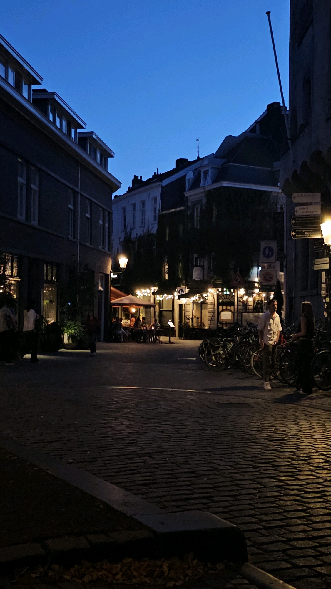 An outdoor evening scene in the city of Maastricht, Netherlands, depicts a cobblestone street lined with buildings. On the left, a dark building with multiple windows is partially visible. In the mid-ground, a restaurant or cafe area features illuminated tables with patrons seated, surrounded by warm string lights and an umbrella. Several bicycles are parked on the right side of the street. Two individuals are standing in conversation near the bicycles, one male and one female. The sky is a deep twilight blue. The overall atmosphere suggests a European city street at dusk.