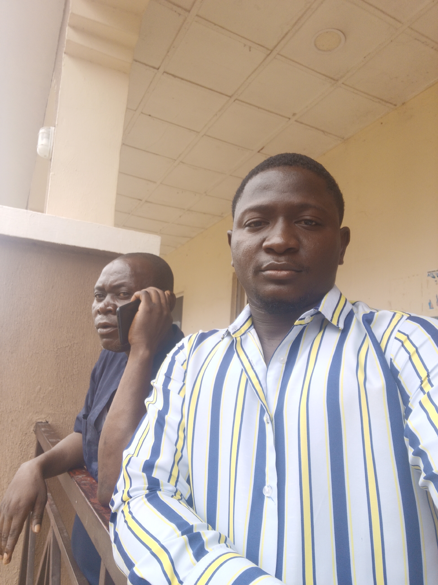 The image features two men of African descent in what appears to be an outdoor or semi-outdoor setting, possibly a balcony or porch, in Akwanga, Nigeria, under natural daylight.
In the foreground, a man with short, dark hair and light facial stubble looks directly into the camera, suggesting a selfie or close-up portrait. He wears a short-sleeved button-up shirt with a distinctive pattern of vertical stripes in white, yellow, and blue. His expression is neutral.
Behind him, slightly out of focus, an older man is captured in profile, engaged in a phone conversation. He wears a dark blue, short-sleeved shirt and is leaning casually on a dark metal railing. He holds a black smartphone to his right ear, and his expression appears serious or thoughtful.
The background consists of light-colored, possibly beige or off-white, building walls and a flat ceiling composed of square tiles. A small, white lighting fixture is mounted on a wall in the upper left. On a wall further in the background, to the right, faint and illegible text or markings are visible. The bright, even lighting indicates it is daytime, and the overall atmosphere is casual.