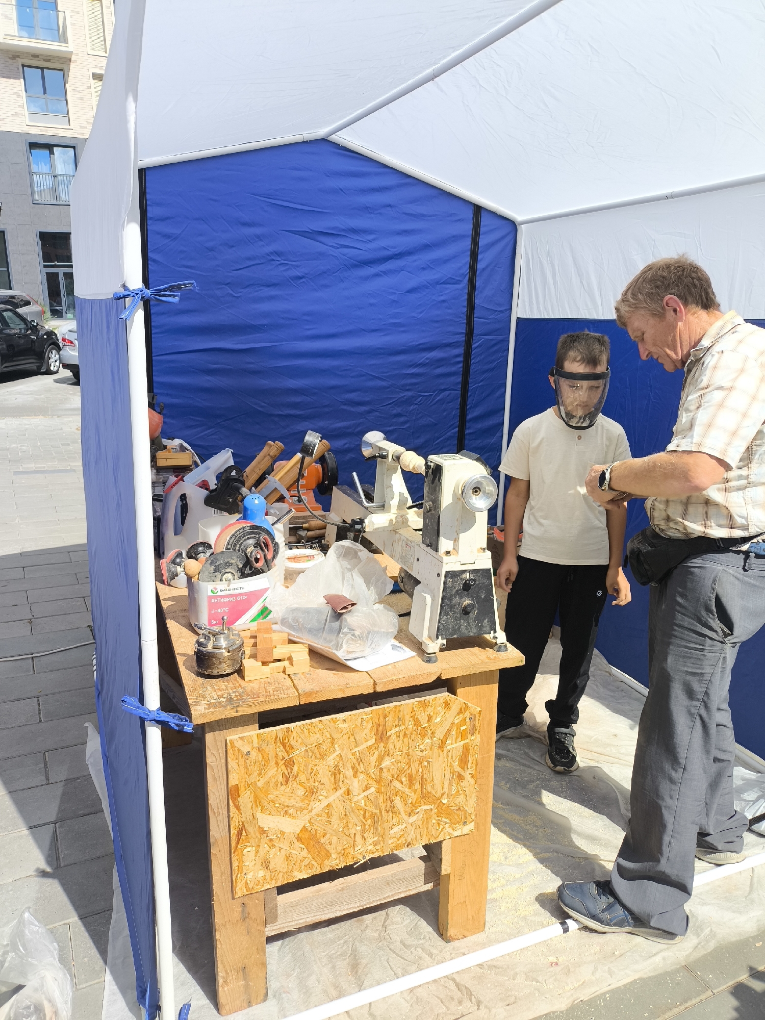 An outdoor demonstration is underway within a blue and white tent structure, possibly at a public event or workshop in Izhevsk, Russia. A man, appearing to be an instructor, is showing a young boy, who is wearing a clear plastic face shield, how to use a woodworking lathe. The lathe is situated on a rustic wooden workbench laden with various tools, including what appear to be chisels, a grinder, and possibly other specialized woodworking implements. Wooden blocks are also visible on the workbench, suggesting an active project. The ground beneath the workbench is covered with a protective sheet, sprinkled with sawdust or wood shavings, indicating recent activity. In the background, a modern brick building with balconies and windows can be seen, characteristic of urban architecture. Cars are parked on a paved area adjacent to the tent.