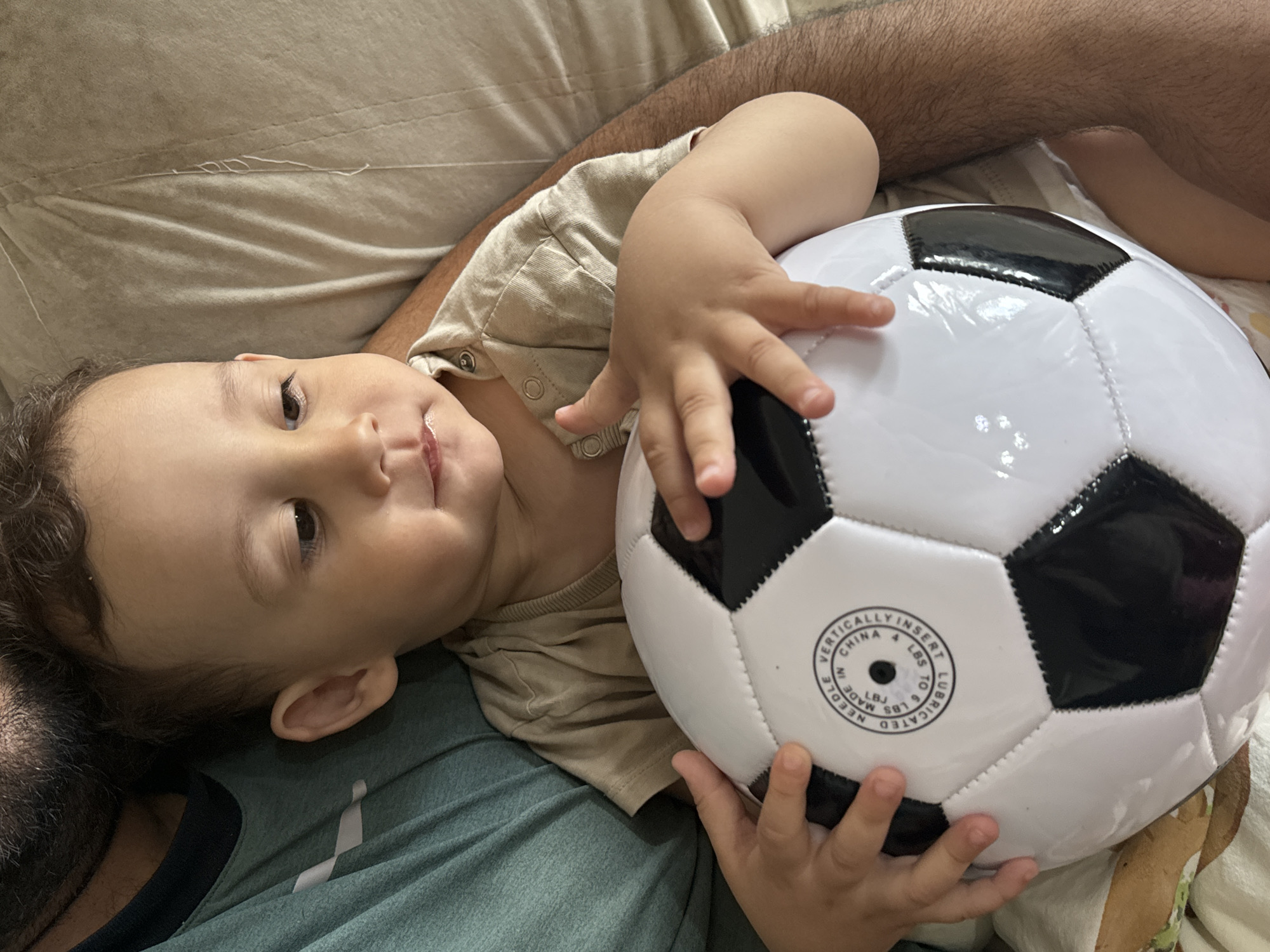 A child is lying down while holding a black and white soccer ball. The child has brown hair and is wearing a beige shirt. The child is lying on top of a person who is wearing a green shirt. The location is unknown, and there is no indication that the image was taken in the city of Chaikovsky, Russia.