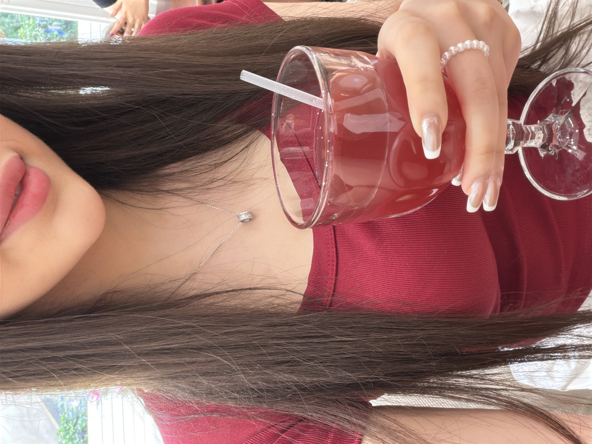 A person wearing a maroon shirt is holding a glass of red liquid with a white straw. The person is wearing a pearl ring on their finger, and they have manicured nails with a white tip. The person also has a silver necklace. The location is unknown, the city of Liangjiang, China cannot be determined from the image.
