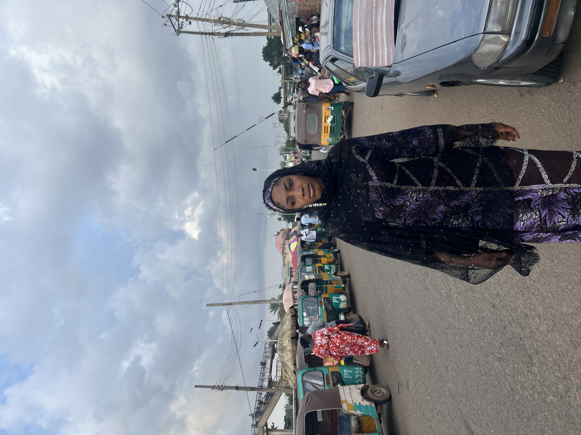 A woman wearing a head covering and patterned garment stands on a street lined with auto rickshaws, likely in Jos, Nigeria. A car is parked on the right, and other people are visible in the background. The sky is overcast.