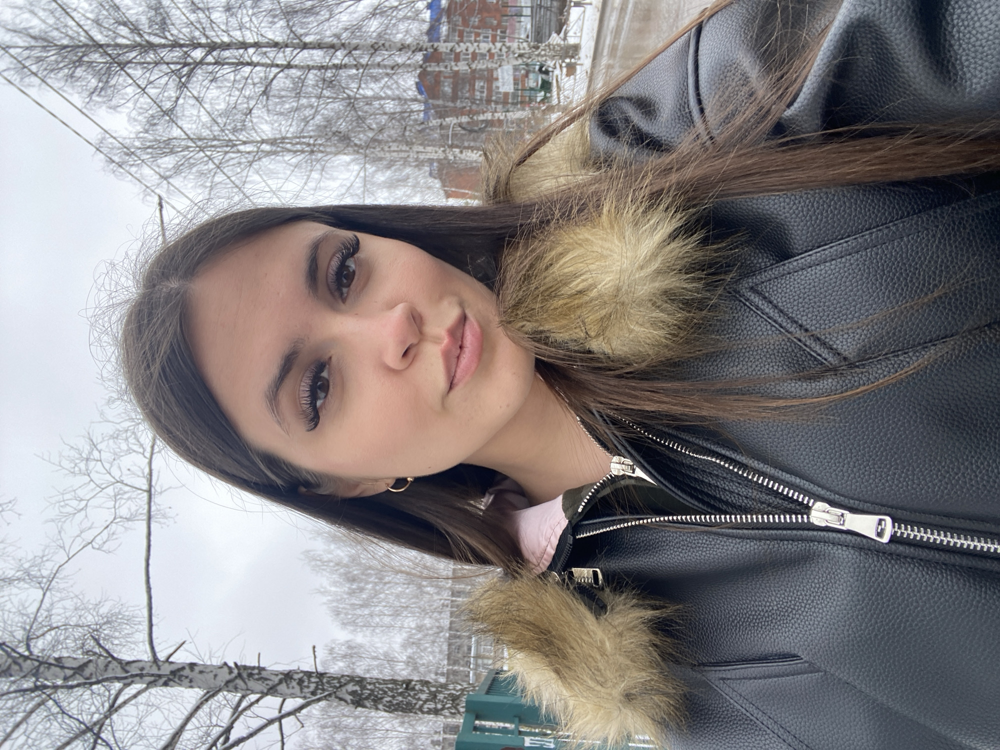 A young woman is taking a selfie outdoors in what appears to be a park or recreational area. She has long, dark hair and is wearing a black leather jacket with a fur-trimmed collar. The background features bare trees, possibly birches, indicative of a winter or early spring scene. In the distance, there are brick apartment buildings, indicating an urban setting. While this does not provide forensic location information, it can be inferred that this location is most likely in the city of Chaikovsky in Russia due to prior information.