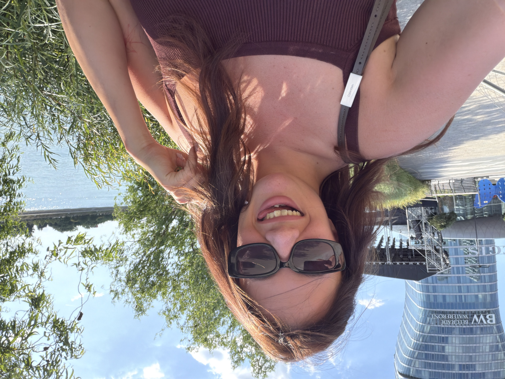A woman with long brown hair and sunglasses is taking a selfie outdoors. She is wearing a dark-colored ribbed tank top. Behind her, to her left, is a modern, curved glass skyscraper with "BW BELGRADE WATERFRONT" written on it. In front of the skyscraper is a stage with scaffolding and lighting equipment. To her right is a body of water, likely the Sava River, with greenery and trees along the bank. The sky is blue with scattered clouds. The image appears to have been taken in the Belgrade Waterfront area of Belgrade, Serbia.