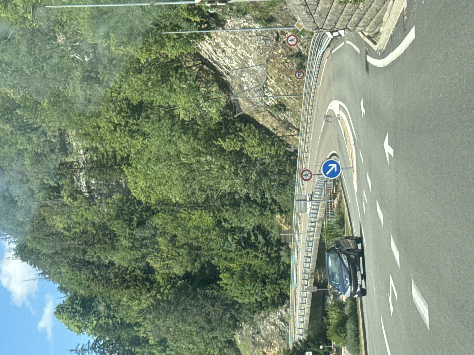 The image captures a road in the city of Hauts de Bienne, France, likely near a mountainous area given the dense evergreen forest covering the hills. The road is paved and marked with white arrows indicating lane directions. In the foreground, a dark colored hatchback car is visible, driving on the road. A concrete bridge spans across the road in the middle ground. Traffic signs are present, including a speed limit sign indicating 70, a pedestrian crossing sign, and a blue circular sign with a white arrow indicating a mandatory turn. The environment appears to be a sunny day with a clear blue sky and some scattered clouds. The road curves to the right, suggesting a winding mountain pass. A stone wall is visible on the right side of the road, and a metal guardrail runs along the edge of the road and the bridge.