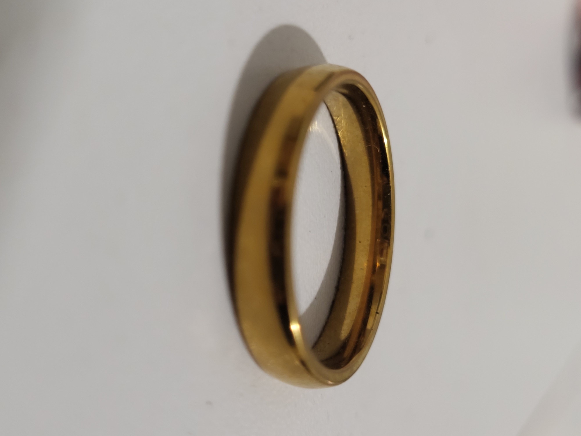 A close-up, side-view shot shows a gold ring positioned on a light-colored, matte surface. The ring is the primary subject, appearing smooth and slightly rounded with a polished finish that reflects the ambient light. A soft shadow is cast by the ring to its left. The background is out of focus and appears to be a plain, neutral color. There are no people, animals, or any indication of a specific location within the city of Horishni Plavni, Ukraine, present in the image. The scene is static and appears to be an indoor product shot.