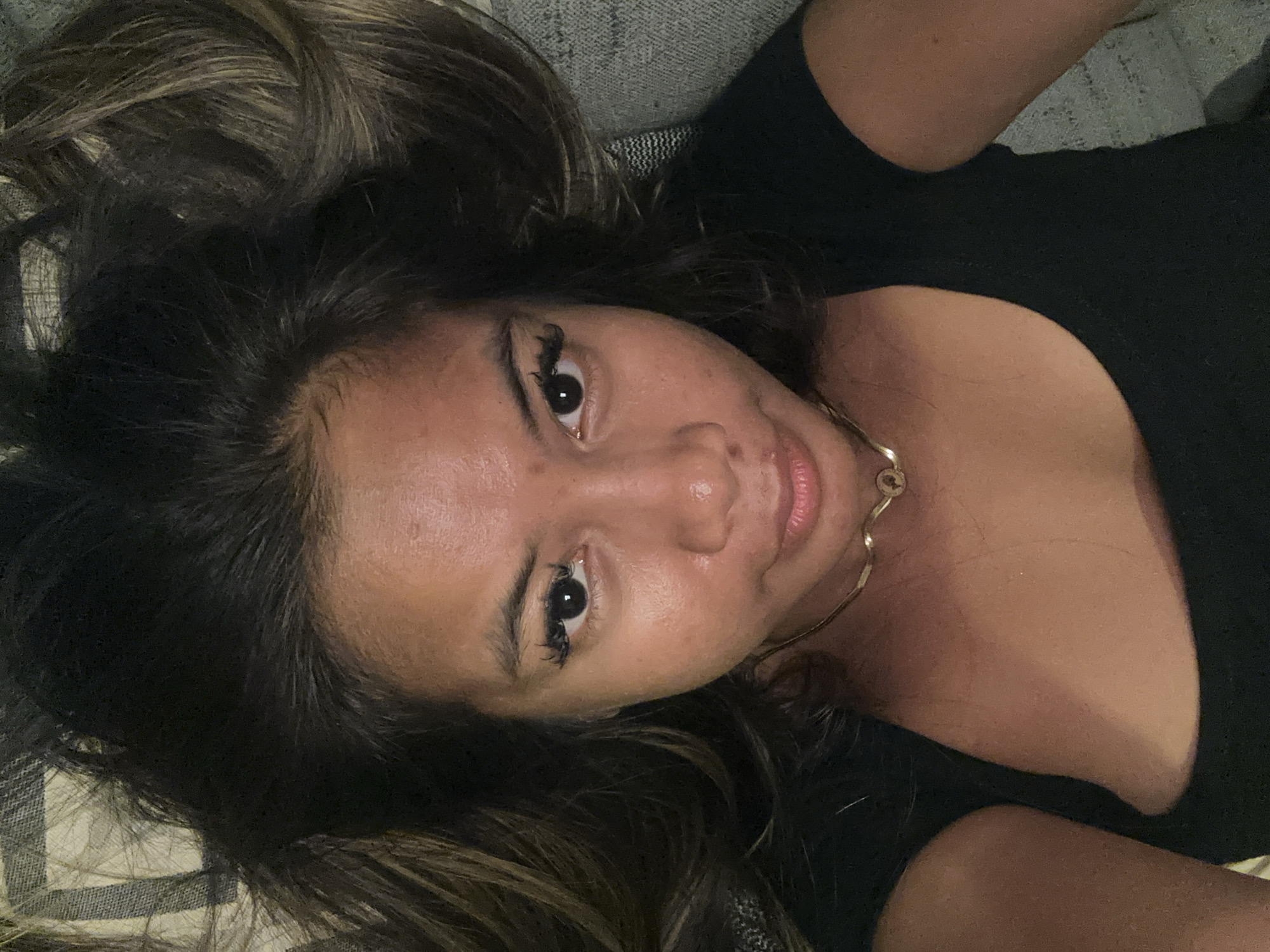 A person is lying down. The person has medium length, dark hair, with some highlights. The person has dark eyes and long eyelashes. They are wearing a black top and a gold necklace. They are lying on a gray and white patterned surface, possibly a couch or bed. It is difficult to determine the location in Central Jakarta, Indonesia from the given image.