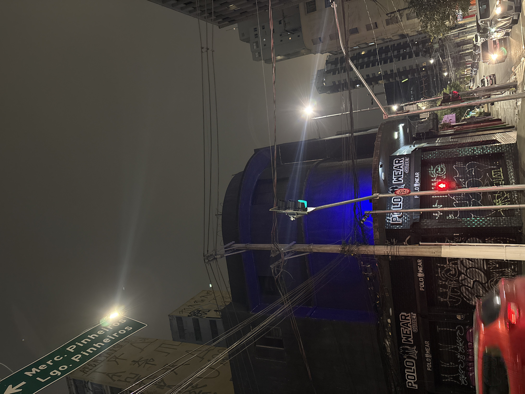 This is a nighttime street scene in the Pinheiros neighborhood of São Paulo, Brazil. A green street sign indicates "Merc. Pinheiros Lgo. Pinheiros" pointing left. A building with a blue illuminated facade is visible, with the storefront below marked "POLO WEAR OUTLET" and displaying graffiti. A traffic light shows a green signal. The street is lined with buildings and overhead electrical wires are abundant. Several cars are on the street, with headlights illuminating the road. The sky is overcast and dark, typical of a city at night.