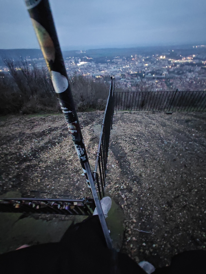 The scene captures a high-angle perspective overlooking the illuminated cityscape of Shumen, Bulgaria, during dusk or early night, consistent with a viewpoint on the Shumen Plateau. The sky above the city is a dark blue-grey.
In the immediate foreground, the lower torso, legs, and light-colored athletic shoes of an individual are visible. The individual's right foot rests near a dark, vertical metal pole, covered in multiple stickers, which runs diagonally from the upper left towards the lower right of the frame. Below this pole, a horizontal metal railing is present, adorned with numerous padlocks, indicating a "love lock" tradition. The ground in the foreground is composed of dark soil mixed with light-colored pebbles and gravel.
A black metal fence, constructed with vertical bars, spans across the midground, delineating the edge of a steep slope. Beyond this fence, the downhill terrain is sparsely covered with barren deciduous trees or bushes. The background features the sprawling city of Shumen, illuminated by numerous scattered lights from buildings and street infrastructure. Faint outlines of distant hills are visible on the horizon, merging with the twilight sky. No specific actions are depicted beyond the static presence of the individual's lower body within the environment.