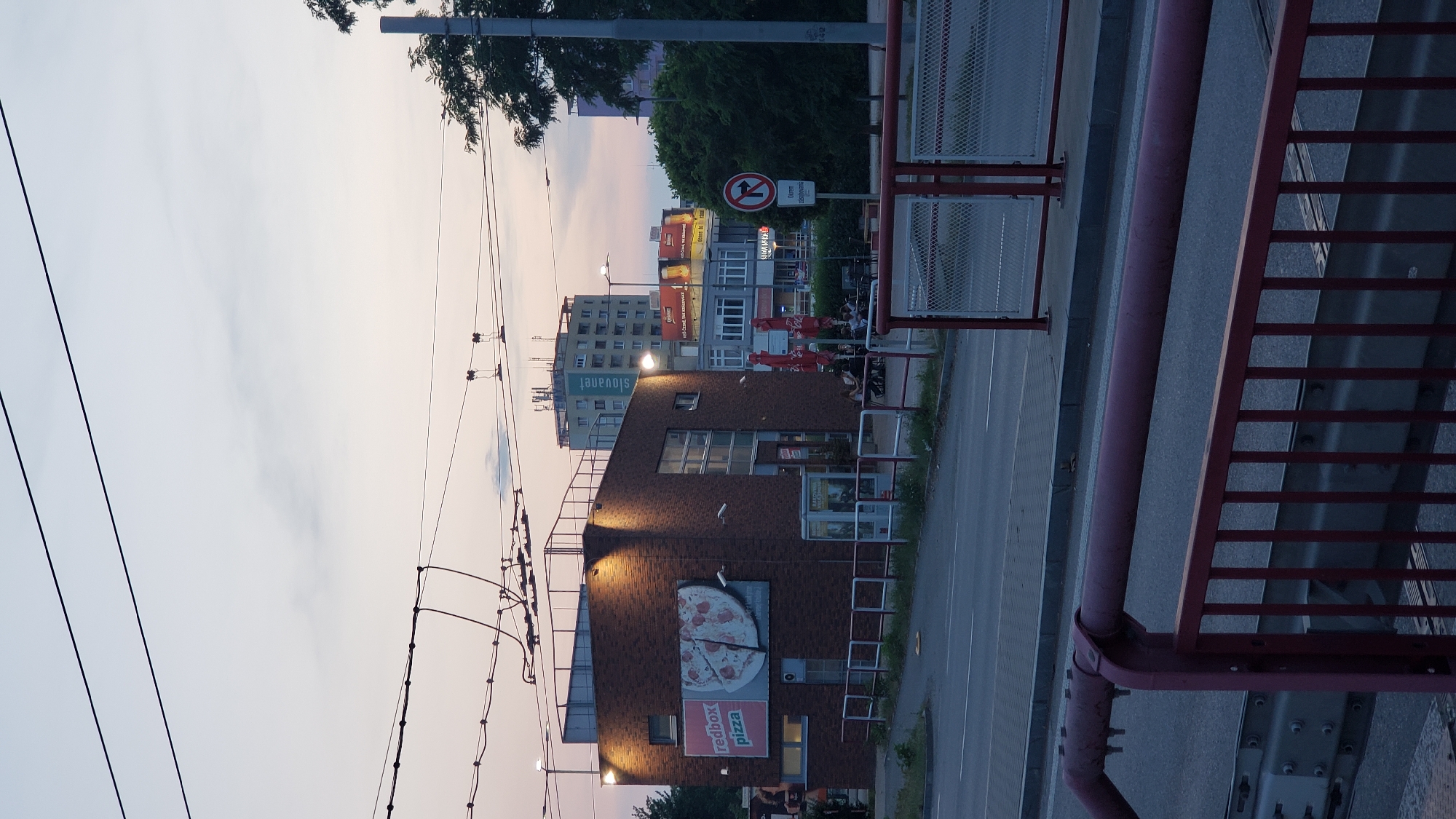 The image captures a street scene in Prešov, Slovakia, during twilight. In the foreground, a red metal railing and fence line a walkway or bridge. Beyond this, a street with tire tracks is visible, with a sidewalk and a redbrick building to the left. This building features a prominent advertisement for "redbox pizza" with an image of a pizza. Several security cameras are mounted on the building's exterior. Further in the background, other buildings are visible, including a multi-story structure with the word "slovanet" displayed prominently. Trees and utility poles with overhead wires are also present. A no-left-turn sign is positioned on the right side of the image. Several people are seated at tables outside the redbrick building, suggesting an outdoor seating area. The sky above is a soft gradient of purple and pale blue, indicative of dusk.
