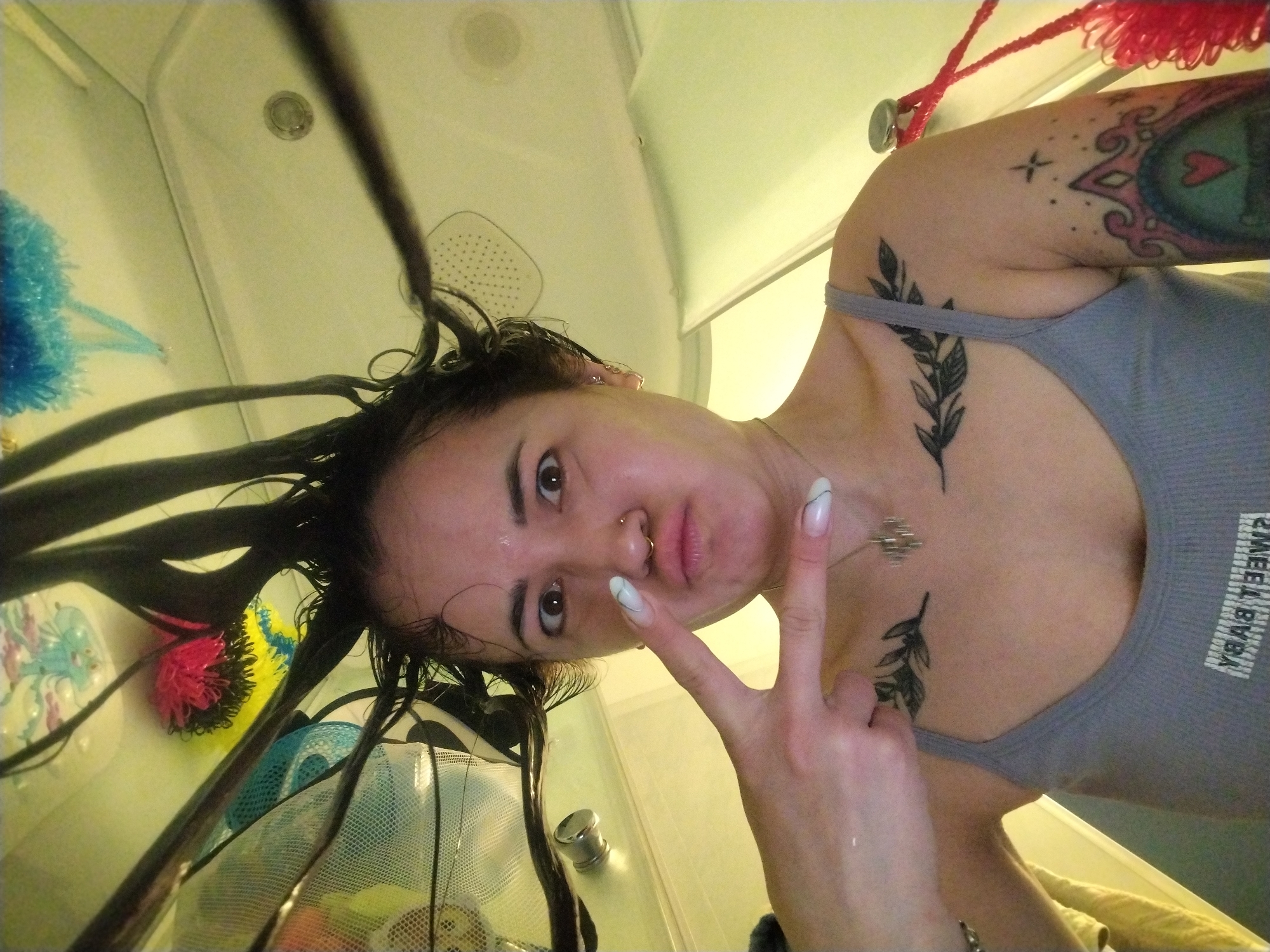 A selfie taken from a low angle of a young woman with wet, dark hair, who is making a peace sign with her right hand. She is wearing a gray tank top. She has a nose ring, and tattoos on her chest and upper arm. The background is a bathroom with a shower. The city of Noviy, Russia is not visible.