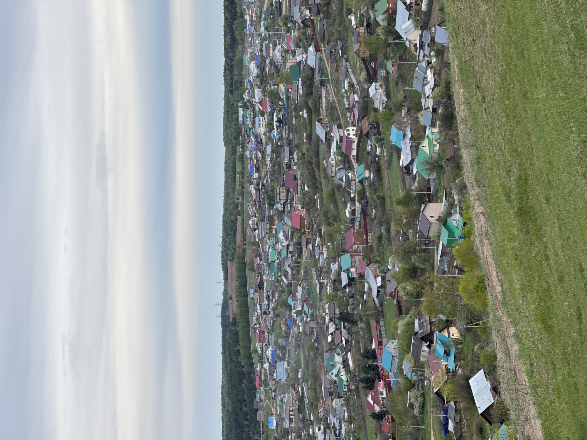 An aerial view of Прикамский, Russia, reveals a dense arrangement of houses with roofs of varying colors, interspersed with trees. A vast green field extends to the right, while a forest lines the left. The sky is overcast, exhibiting shades of white and light blue.