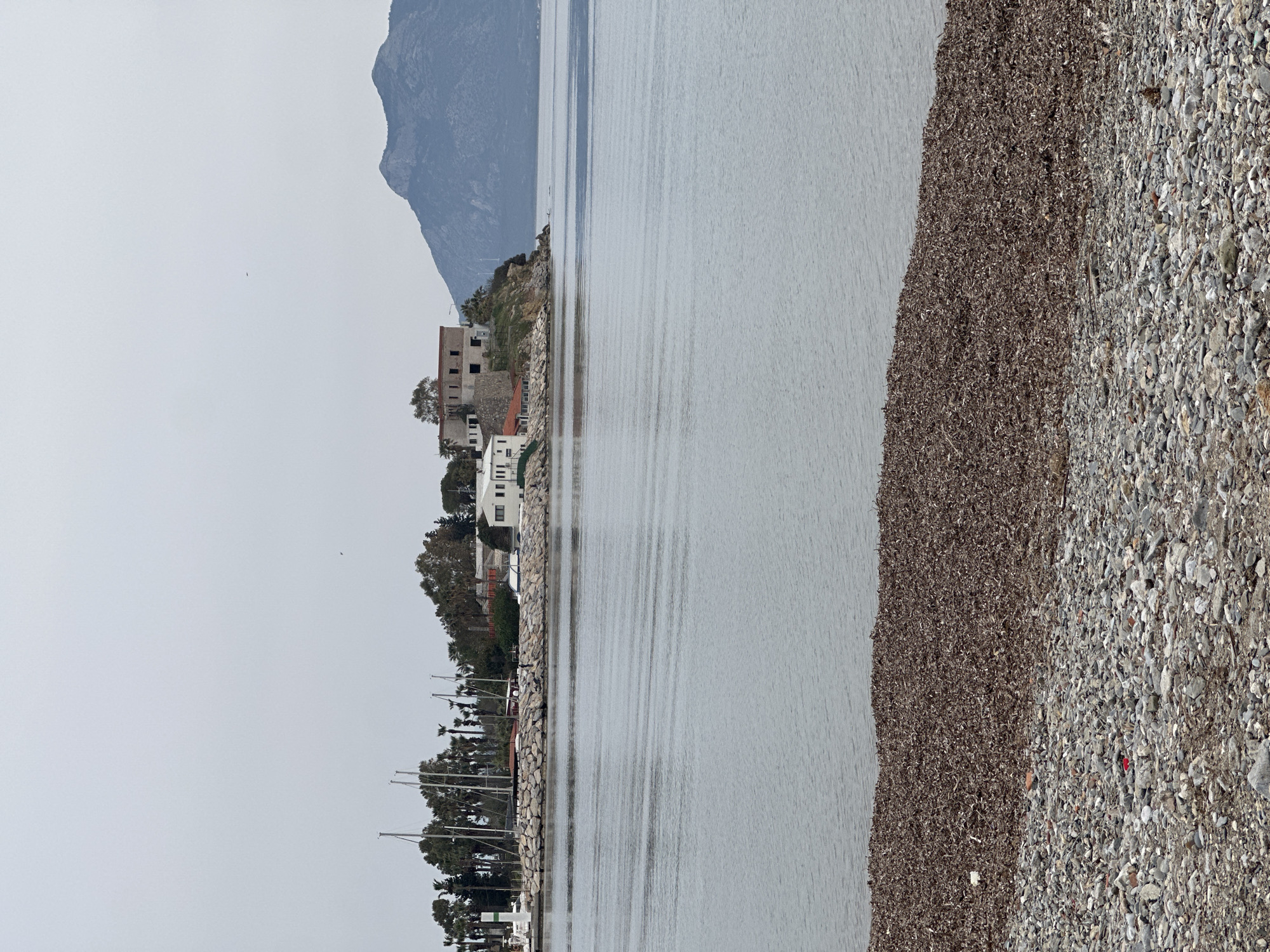 The image depicts a view from the waterfront in Datça, Turkey. A pebble beach lines the right side of the frame, with a darker band of seaweed separating it from the grey water. On the opposite shore, there is a rocky outcrop with vegetation and several white buildings. A hill rises in the background. The water is calm, with subtle ripples. Along the left edge of the frame, there are trees and the masts of sailboats are visible. The sky is overcast and grey. A single bird is visible in the sky.