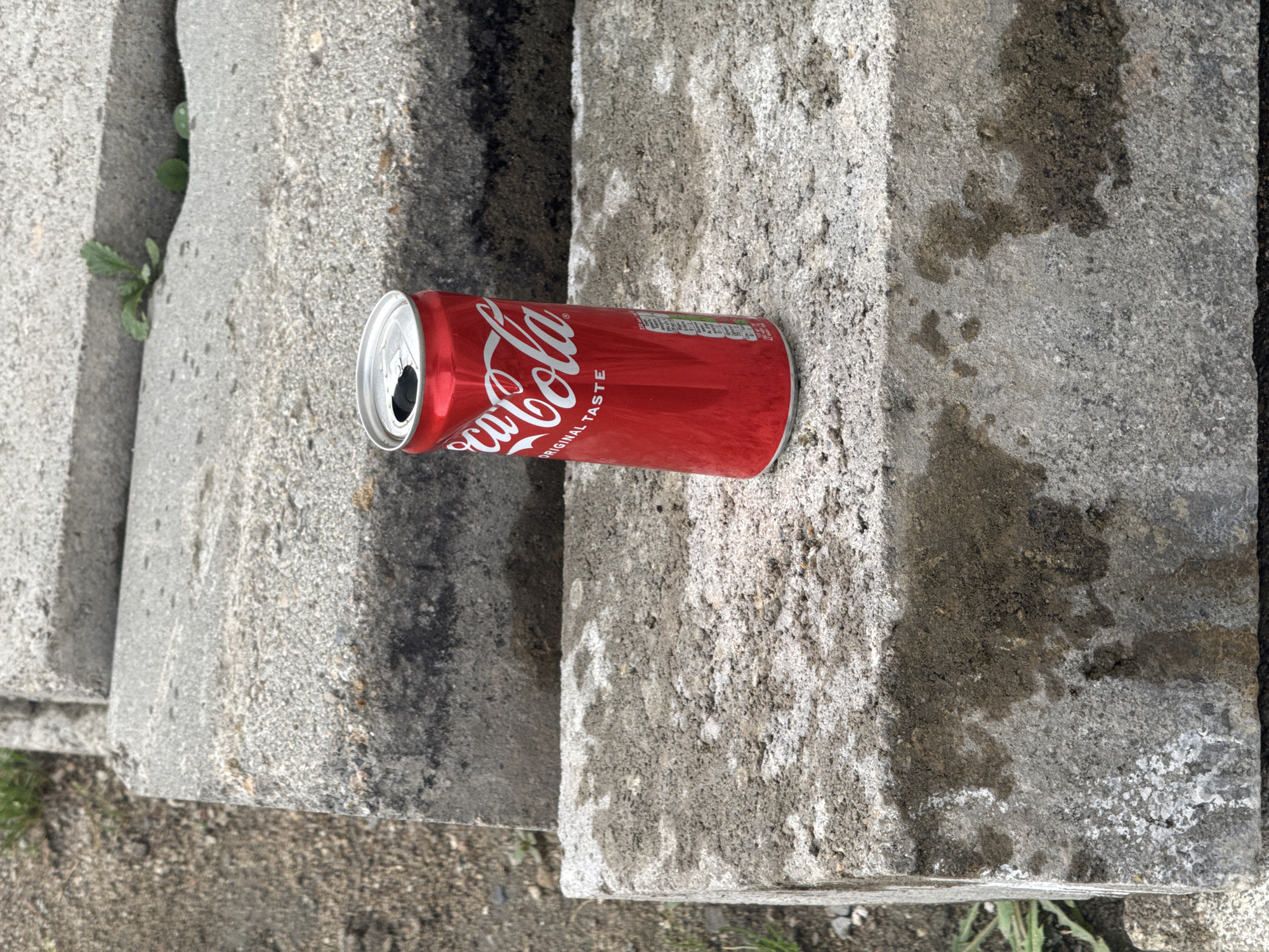 An eye-level outdoor shot depicts a red Coca-Cola can lying horizontally on a weathered, grey concrete surface in Kalekovets, Bulgaria. The can is open, with its top rim appearing slightly crumpled, and it seems to be empty or partially consumed. The distinctive white "Coca-Cola" logo in its script font is prominent on the can's side, along with the text "ORIGINAL TASTE." Faint nutritional information is also visible on the side of the can.
The concrete surface is composed of large, rough-textured blocks or slabs, displaying signs of age, dirt, and dark moisture stains, particularly on the block to the right of the can. Small green plants and weeds are growing in the crevices between the concrete blocks and in the dirt or gravel ground visible in the background and foreground, suggesting an untended outdoor environment. The scene is illuminated by natural daylight, without strong shadows indicating a specific time of day beyond daytime. The image captures a static moment, focusing on the discarded can within this somewhat rugged, utilitarian outdoor setting.