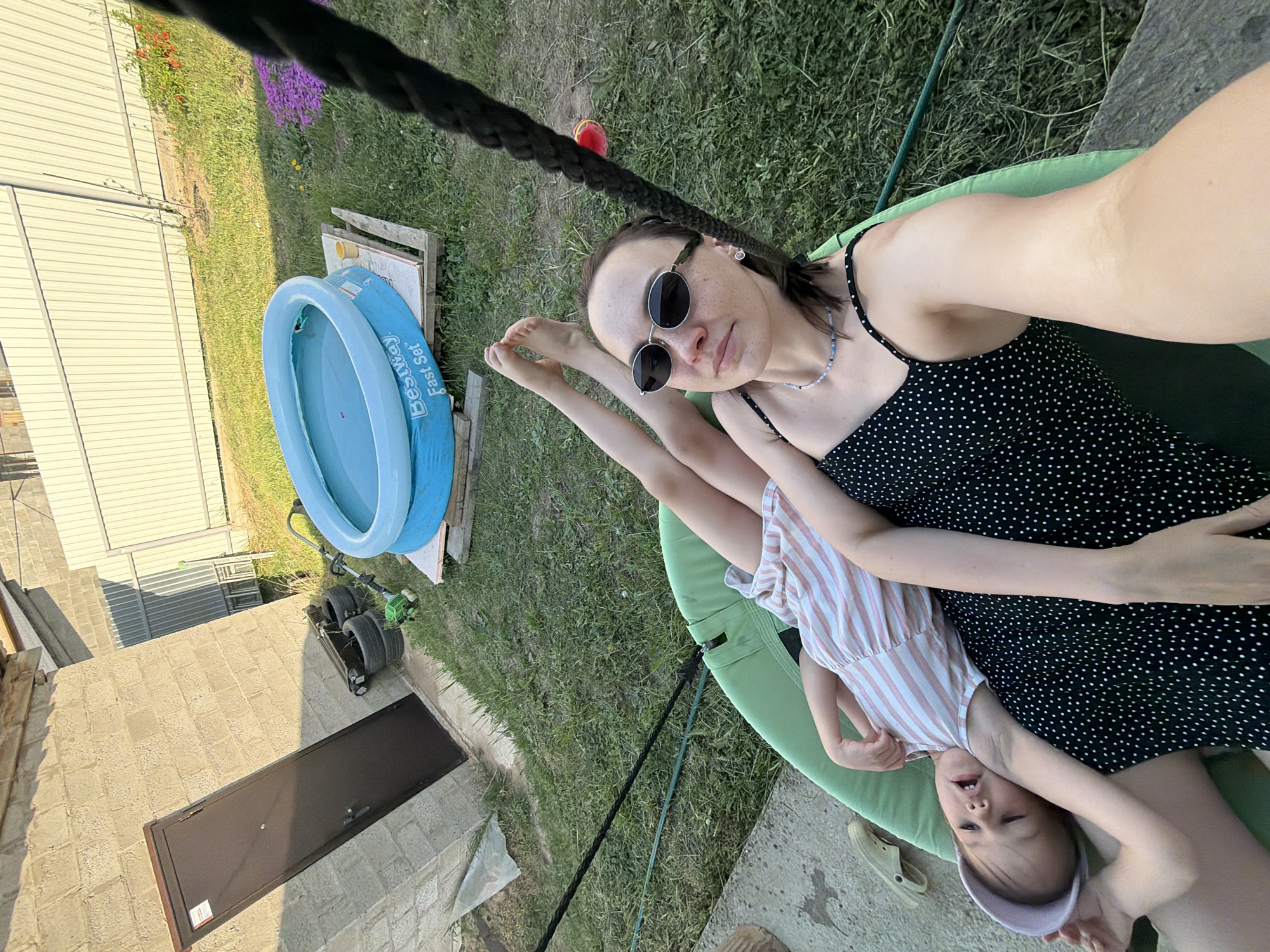 The image shows a fair-skinned woman wearing a black dress with white polka dots and sunglasses, reclining in a green hammock chair with a child in a striped dress. The child is upside down. The location appears to be an outdoor residential area in Chaikovsky, Russia. A blue inflatable pool rests on a wooden pallet next to a white metal shed. There's a dark grey door in a cement base structure. A black rope is visible, likely part of the hammock's suspension. Green grass covers the ground.