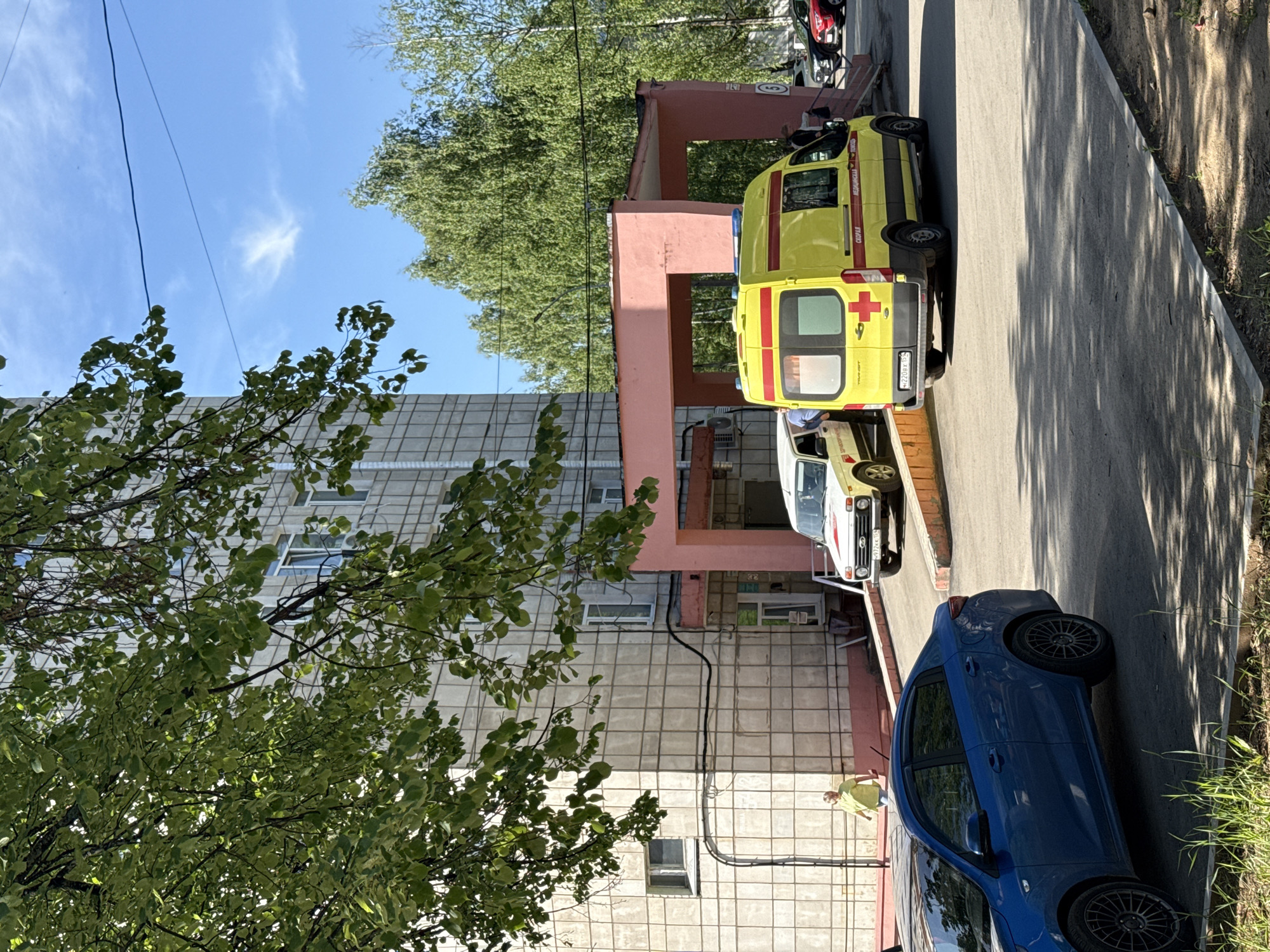The image shows a street scene in Chaikovsky, Russia, with a row of vehicles parked along a sidewalk next to a building. From left to right, a blue sedan, a white automobile, and a yellow ambulance are parked near the building's entrance. A figure wearing a yellow shirt and dark-colored pants stands near the building's exterior wall. A tree is visible to the left, partially obscuring the building. The building has a pink entrance structure. The sky is blue with some clouds visible.