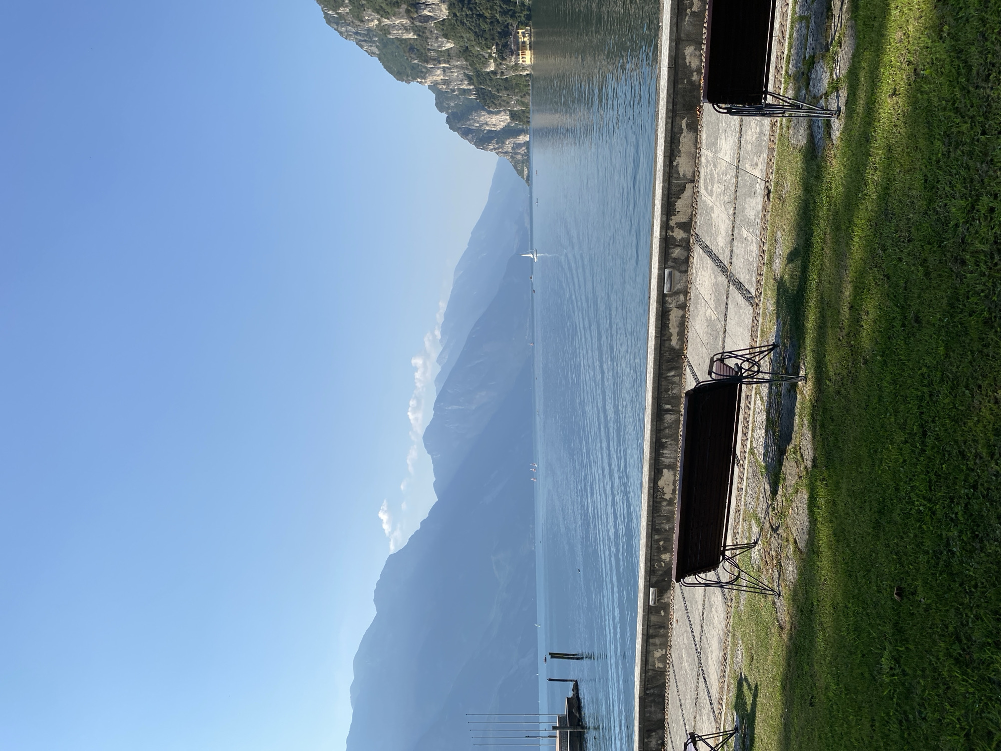 The image depicts a serene lakeside scene in Riva del Garda, Italy. In the foreground, two dark-colored benches with metal frames are situated on a manicured lawn, casting shadows on the grass and paved walkway behind them. The walkway, made of stone tiles and pebbles, runs along the edge of a calm, blue lake. Beyond the lake, a series of majestic mountains rise in layers, their peaks fading into a hazy blue sky. Several sailboats are visible on the water, adding a touch of activity to the tranquil landscape. On the right side of the frame, a steep, rocky mountainside covered in lush green vegetation slopes down to the lake. A distinct yellow building with a red roof is nestled at the base of this mountainside, overlooking the water. The clear, bright sky suggests a sunny day, contributing to the overall peaceful and picturesque atmosphere of the location.