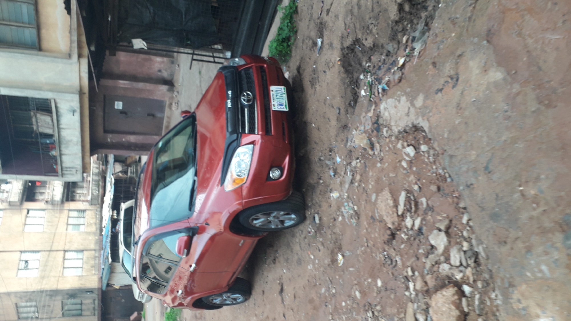 The image depicts a red Toyota RAV4 parked on a muddy street in what appears to be Onitsha, Nigeria. The environment is littered with trash and debris, suggesting a lower-income or neglected area. To the left of the car is a faded, multi-story building with visible windows. A narrow alleyway is seen next to the building. The car has a Nigerian license plate with the registration "BMR 172DJ". The composition is somewhat tilted, and the overall impression is one of urban decay or neglect.
