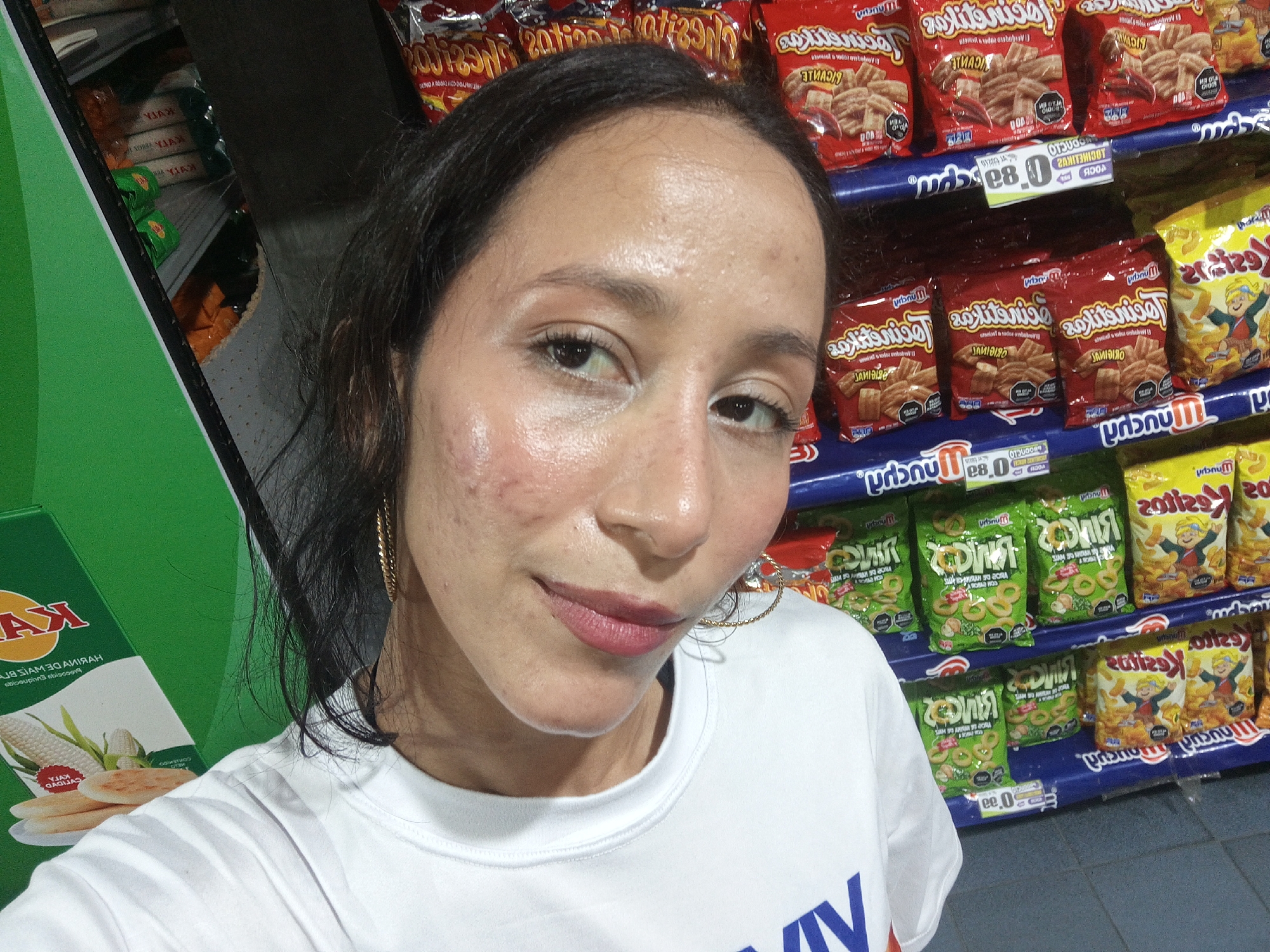 A woman stands in front of a store shelf stocked with snack foods, likely in a supermarket. The snacks include different flavors of "Locinelikuz" and "Kysitos" brands of potato chips and rings. The woman wears a white t-shirt and gold hoop earrings. Her hair is dark and pulled back, and she is wearing makeup. In the background on the left, there is a green shelf with "Kyla" brand corn flour visible. The price tags indicate the location is most likely Porlamar, Venezuela.
