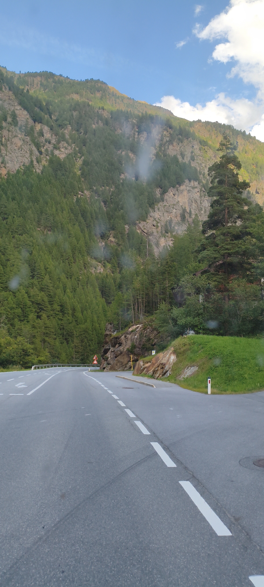 The image shows a road winding through a mountainous landscape. The road is asphalt with white dashed and solid lines indicating lanes. On the left, the road curves to the left and is bordered by a steep, forested mountainside. On the right, there is a grassy embankment with a rock face. The mountains are covered in dense green pine trees, and the sky above is blue with some white clouds. There are a few small plumes of mist or smoke visible on the mountainside. A red and white triangular warning sign is visible on the left side of the road, and a white marker post is visible on the right. The overall scene suggests a scenic drive through a mountainous region, possibly in the Austrian Alps, given the context of Sölden, Austria.