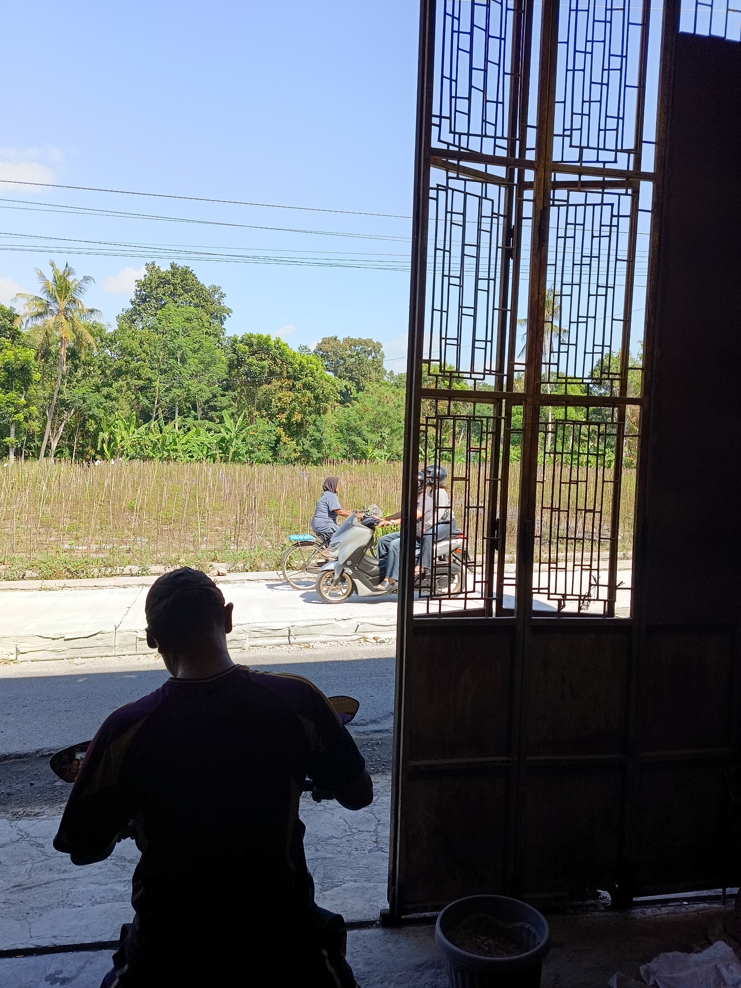 The image captures a scene on a bright, sunny day in what appears to be a rural or semi-rural area of Sukorini, Indonesia. In the foreground, a person is silhouetted, seated on a motorcycle facing away from the camera. Their back and the back of their head are visible, wearing a dark-colored shirt with a purple accent. The motorcycle is also mostly in shadow.
Through a large, ornate metal gate, the background reveals a street with two individuals on a motorcycle. One person, wearing a helmet and a gray long-sleeved shirt, is driving, while another person is a passenger behind them, also wearing a helmet and a dark-colored top. To the left of the street, there is a paved walkway, and beyond that, a field with dry stalks or reeds. Further back, a dense line of lush green trees forms the horizon, with a few taller palm trees interspersed. The sky is a clear, bright blue. The gate itself is made of dark metal, with a repeating pattern of rectangular openings. To the far right, a dark interior space is visible, with a grey bucket in the lower right corner.