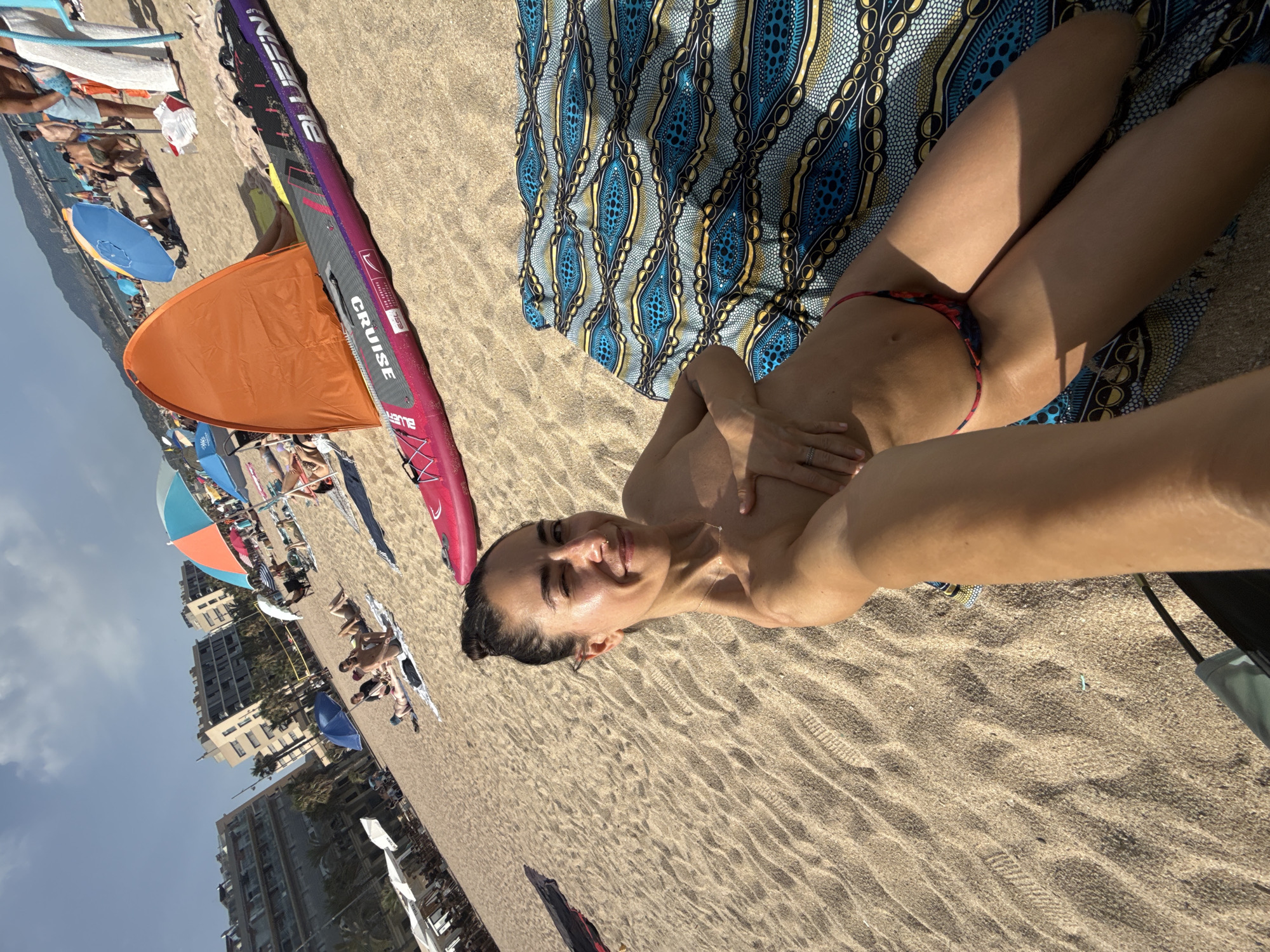 A woman is lying on a patterned blanket on a sandy beach, with her arm extended as if taking a selfie. She is wearing a small bikini bottom and her chest is covered by her hand. The beach is populated with other people sunbathing and relaxing under umbrellas. In the background, there are buildings, trees, and the sea. The image captures a sunny day at the beach in Badalona, Spain, with the distinctive architecture of the city visible. A paddleboard and an orange beach tent are also present in the scene.