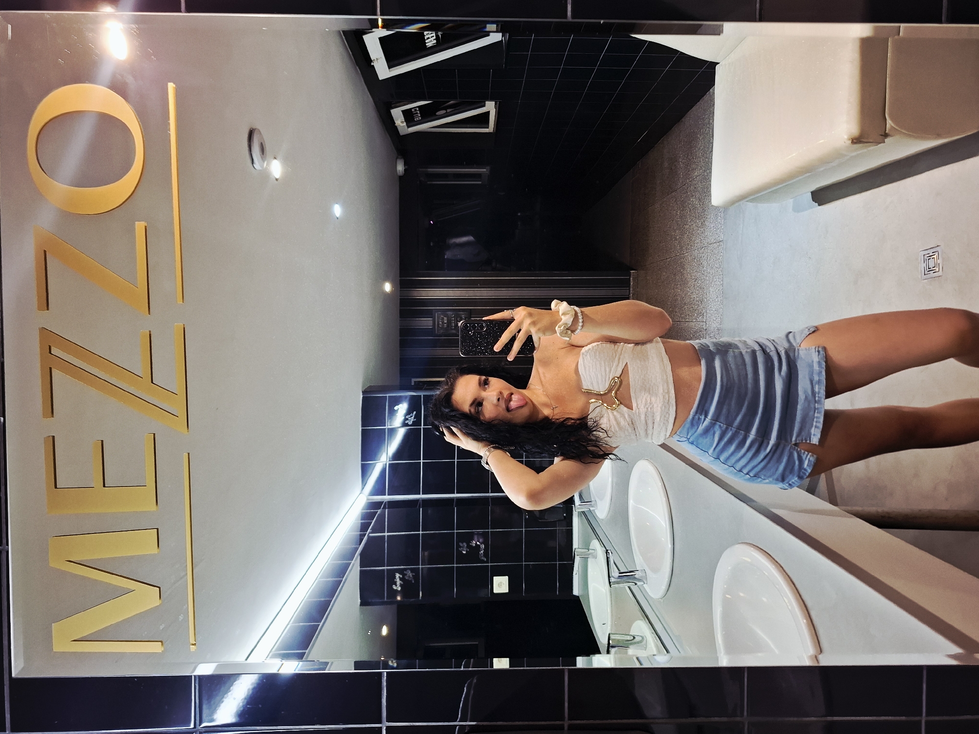 The image is a mirror selfie taken in a dimly lit, modern bathroom. The woman in the photo is in the foreground, her reflection dominating the frame. She has dark, curly hair, is sticking her tongue out, and is touching her hair with her right hand. She is wearing a white strapless top with a gold embellishment and a light blue denim skirt with a thigh-high slit. Her left hand is holding a smartphone, which is capturing the image.
The bathroom features black tiled walls and a long, light-colored countertop with multiple sinks. The word "MEZZO" is prominently displayed in gold lettering on the mirror above the sinks. In the background, a hallway is visible, with framed art on the wall and a white bench or seating area on the right. The overall aesthetic suggests a trendy establishment, possibly a club or a high-end establishment. The image does not provide any specific information about the city of Mezdra, Bulgaria, or any connection to it.
