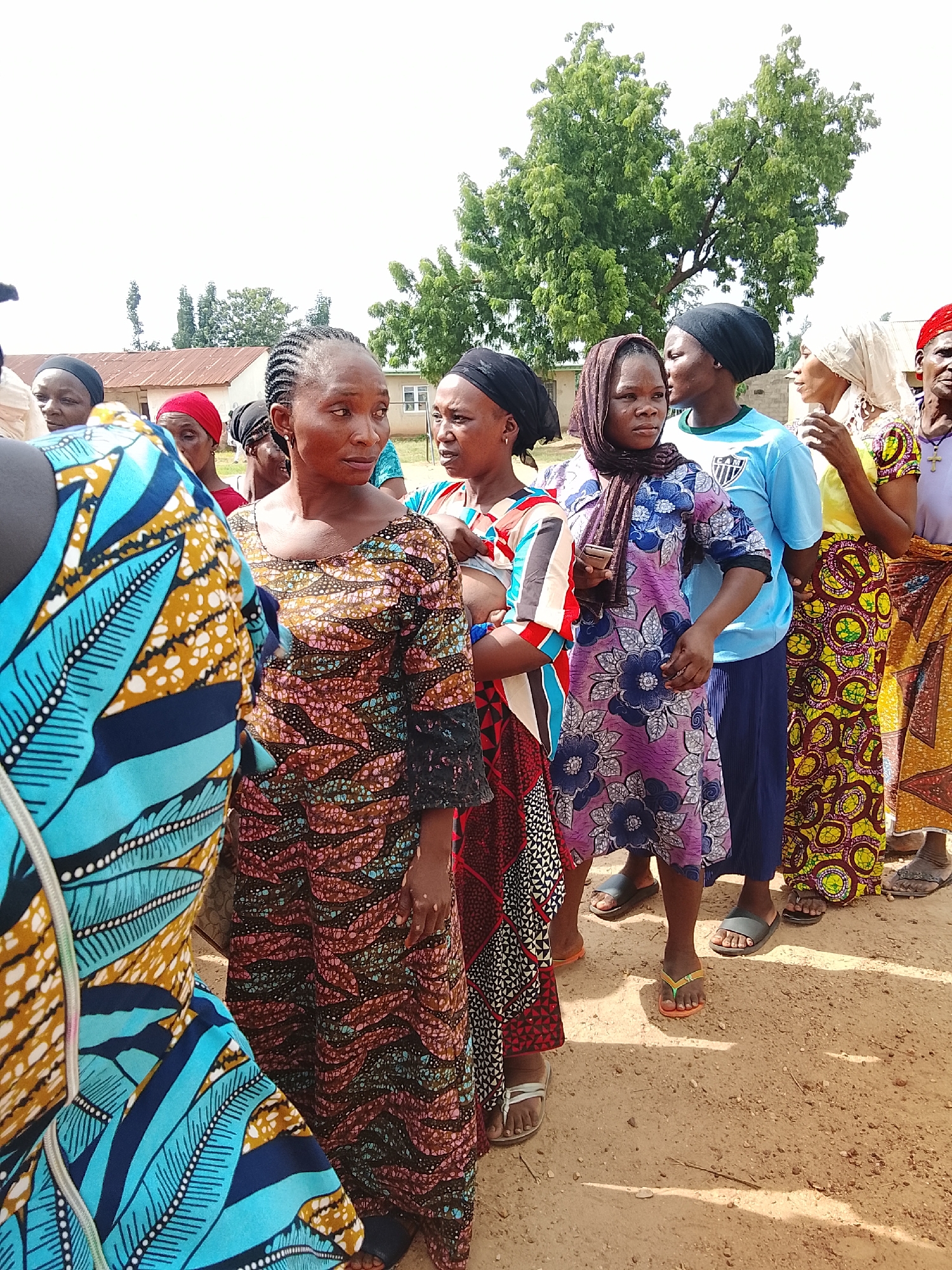 A group of women are standing in a line outdoors, seemingly waiting for something. They are dressed in brightly colored traditional African attire, with vibrant patterns on their dresses and headscarves. Some of the women are looking towards the camera, while others are looking in the same direction as the line. The setting appears to be a rural area, with a dirt path, some trees, and simple buildings in the background. The sky is bright and clear, suggesting it is daytime. The overall mood seems to be one of quiet anticipation. There is no visible text in the image.