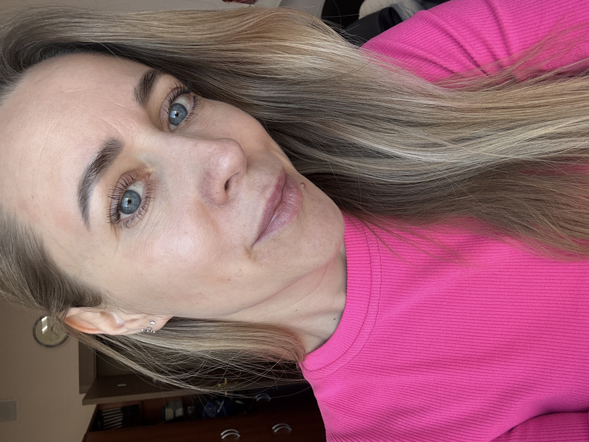 A close-up portrait, likely a selfie, features a woman indoors in Chaikovsky, Russia. She has long, straight, ash blonde hair that flows around her. Her blue eyes are prominent, enhanced by dark, well-defined eyelashes and eyebrows. Her fair skin shows natural texture, with a small mole visible above her upper lip on the right side. Her expression is neutral and calm as she looks towards the camera. She wears a vibrant hot pink ribbed long-sleeved top. In her visible left ear, she has multiple small silver earrings, including two hoops with tiny stones. The background is softly blurred but reveals an indoor setting with a partial view of a round wall clock with a light face and dark hands, along with some dark furniture below it. The bright, even lighting suggests it is daytime. No text is visible in the image.