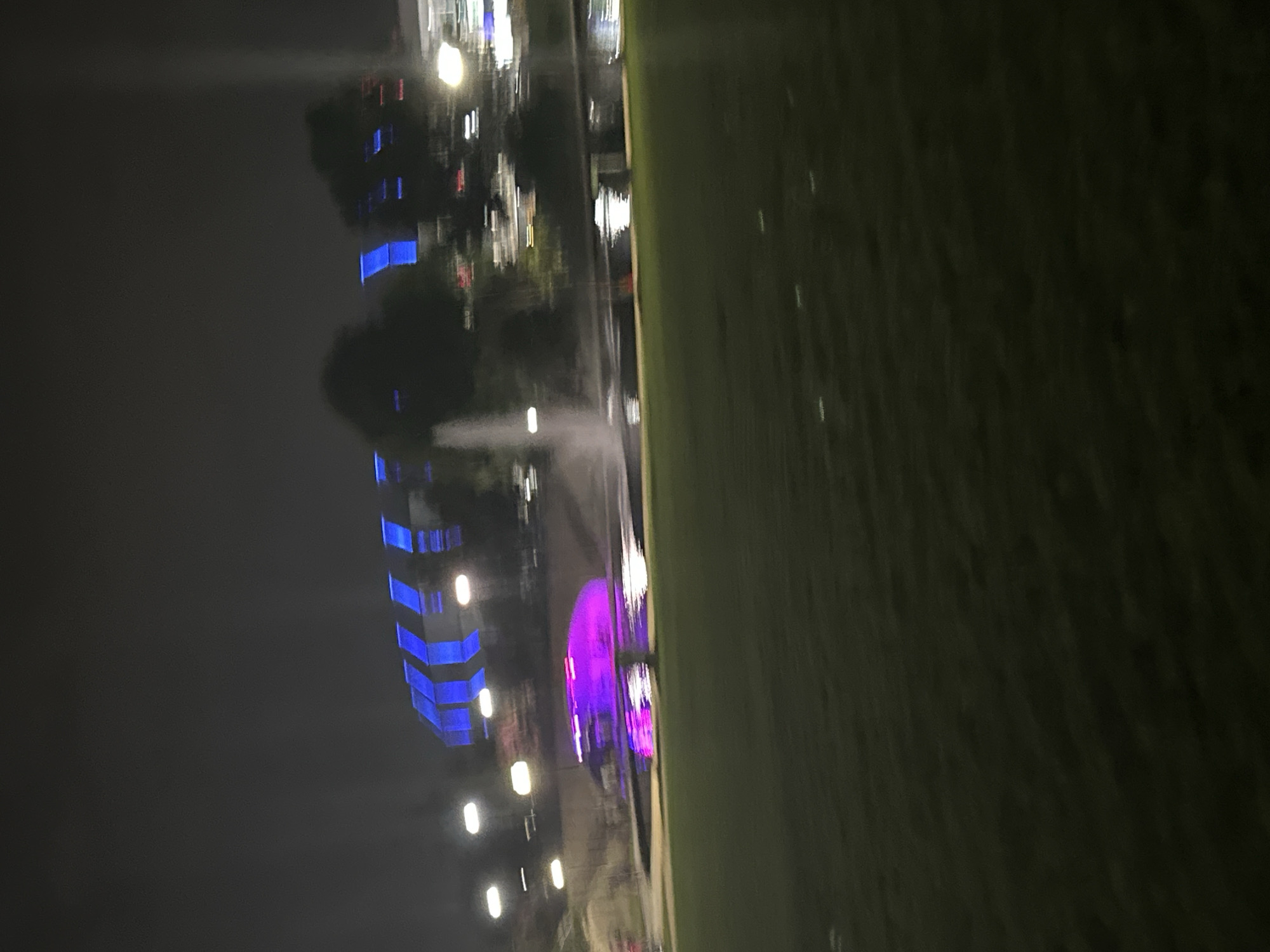 The image depicts a night scene with a blurred perspective, suggesting motion. In the background, a structure with vibrant blue, angled lighting is visible. To the left of this structure, a fountain sprays water, illuminated by a strong pink and purple light source originating from within a shallow body of water. The foreground consists of a dark, grassy area. The scene is located at Big Spring International Park in Huntsville, Alabama, United States of America.