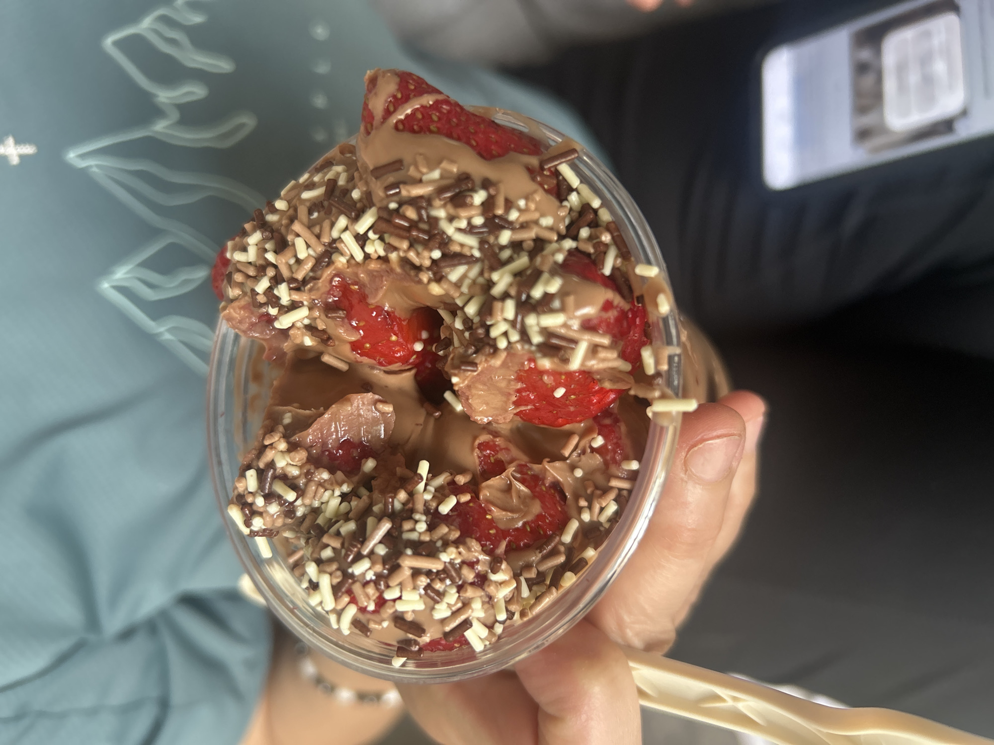 A close-up, high-angle shot depicts a plastic cup filled with strawberries, chocolate sauce, and sprinkles. The cup is being held in a person's hand. The background includes a person wearing a light blue shirt on the left and another person holding a cell phone on the right. The location is listed as Unknown, Unknown.