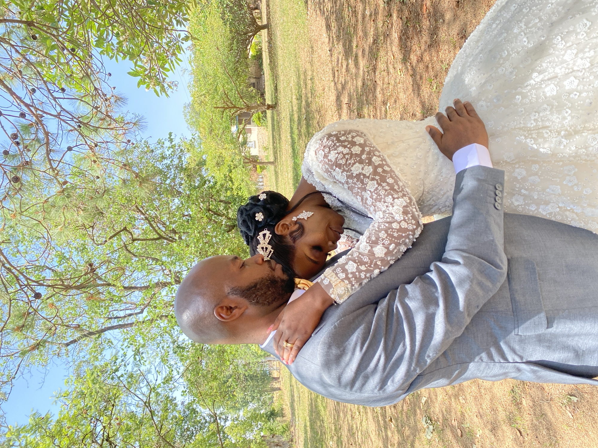 In Dutsen Kato, Nigeria, a newlywed couple is outdoors in what appears to be a park or garden-like setting. The bride, wearing a white, long-sleeved lace wedding dress with elaborate hair accessories, is embraced by the groom, who is in a grey suit. The groom's head is bald, and he has a neatly trimmed beard. The couple is tightly embracing, their faces close, conveying affection. The location is characterized by grassy ground, trees, and clear blue sky. The sunlight filters through the trees. The setting appears to be tranquil and celebratory.