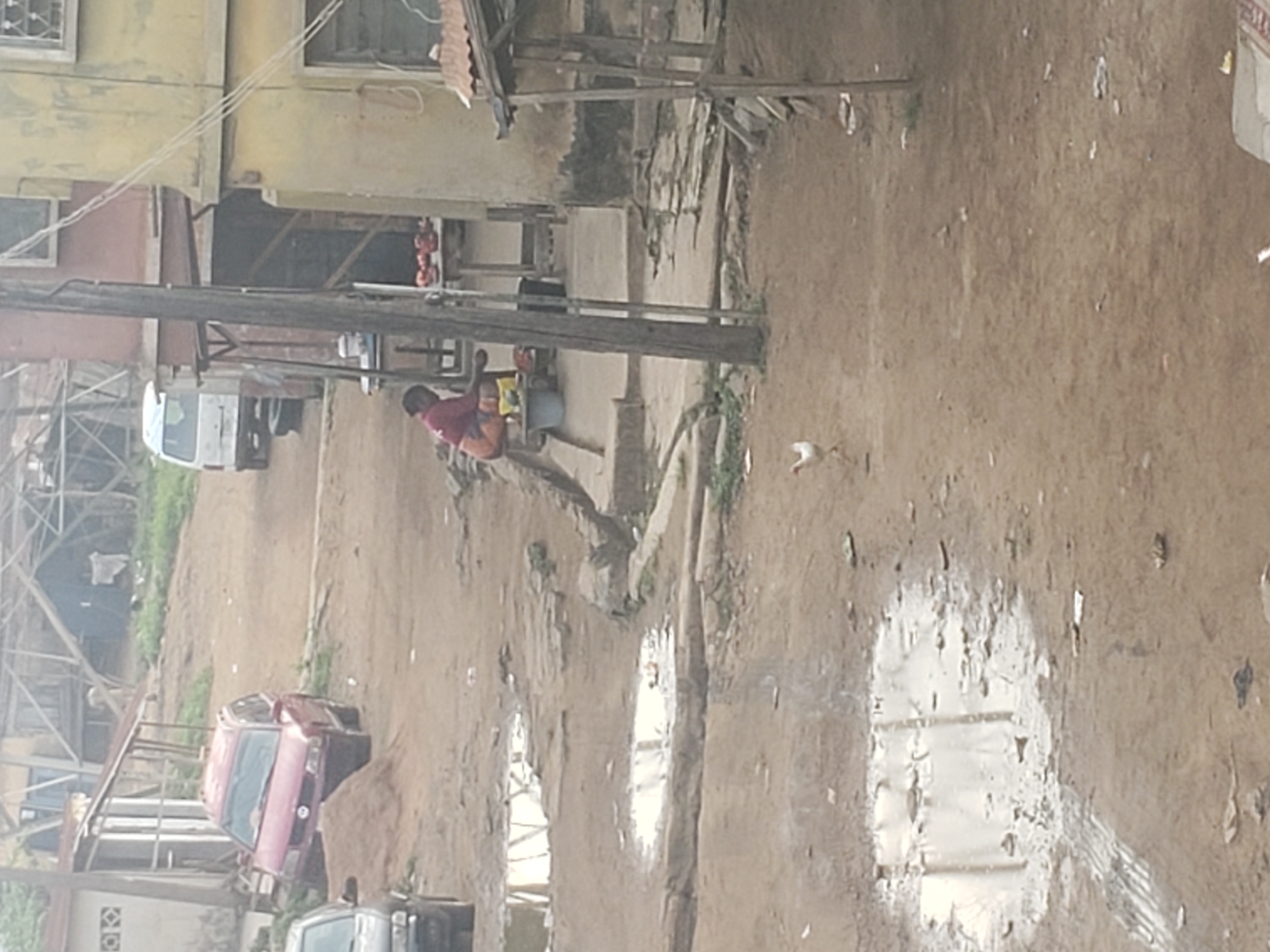 The image shows a street scene in Alalubosa, Nigeria. The road is unpaved and muddy with puddles reflecting the sky. A woman wearing a red top and patterned skirt is bending over a bucket near a building. A small wooden structure with displayed goods sits next to her. Vehicles, including a white van and a red car, are parked along the side of the road. Buildings of varying construction are present, some with wooden frames and others with more substantial walls. Power lines stretch across the scene.