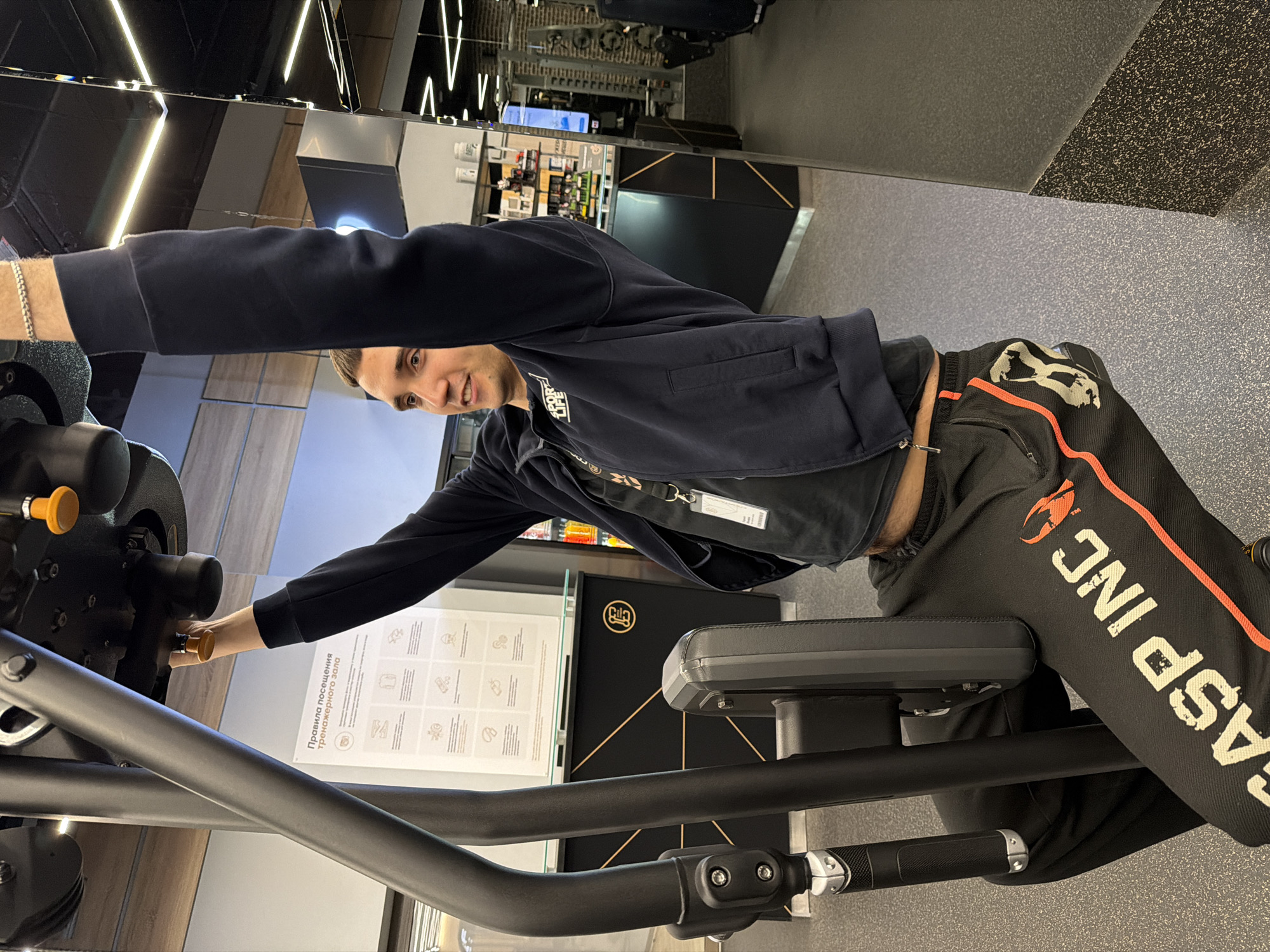 A man is at a gym, adjusting a black exercise machine. He is wearing a navy blue zip-up jacket and black sweatpants with the word "GASP INC." printed in white on the right leg. The background features gym equipment and signage with Russian text. The location is in Saint Petersburg, Russia, based on the presence of the gym "Rila" visible behind the exercise machine.