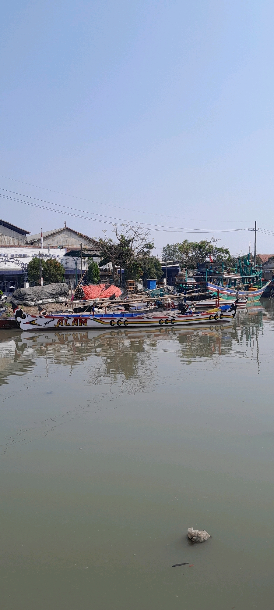 A wide shot captures a murky canal in Pasuruan, Indonesia, lined with docked fishing boats and a commercial building. The sky is a clear, pale blue. The canal's surface is calm, reflecting the boats and the overcast sky. A few pieces of debris float on the water, including a plastic bag on the right side.
In the foreground, several colorful fishing boats are moored side-by-side. The closest boat has a white hull with red, yellow, and blue stripes along its length. It is emblazoned with the word "ALAM" in red lettering and "BERKAH" in smaller red text above it, along with multiple instances of "CCC" in red. The boat is covered in tarps and has a decorative black and white design on its bow. Other boats are visible behind it, displaying similar colorful designs.
Behind the boats, a large industrial-style building dominates the background. Its facade is light-colored with a sloped roof and several windows. A red and white flag flies from a pole in front of the building. Trees with sparse foliage are scattered around the building and along the canal's edge. Electrical wires crisscross the scene, supported by utility poles. The overall impression is one of a working port or industrial area.