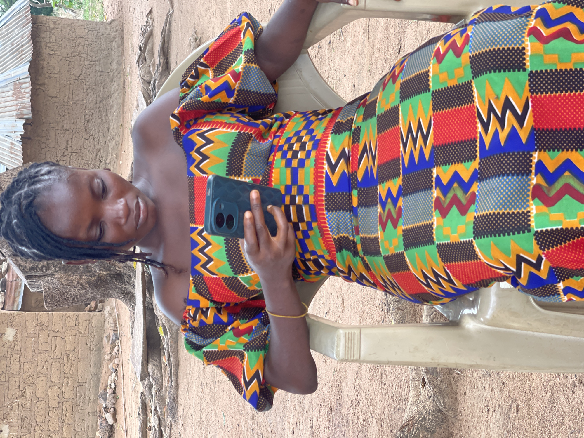 A young Black woman is depicted in an outdoor setting, seated and engrossed in her smartphone. Her hair is styled in short dreadlocks or twists, and she has a calm, focused expression as she looks down at the device. She is wearing a vibrant, floor-length dress adorned with a complex, multi-colored geometric pattern, typical of West African textiles, featuring squares, zigzags, and dots in hues of red, green, blue, yellow, and black. A thin, yellow bracelet is visible on her right wrist.
The woman holds a dark-colored smartphone with her right hand, showcasing a distinct triple-camera module on its back. She is seated on a light-colored, possibly beige or off-white, plastic chair.
The background reveals a simple, earthy environment, consistent with a rural or semi-urban area in Tabu, Nigeria. To her left, a structure with corrugated metal sheeting (likely a roof or wall) stands beside a textured wall made of mud or plaster. The ground beneath her and extending into the background is dry, earthy soil with visible small rocks and roots. The bright, even lighting suggests daytime, likely mid-morning or afternoon, under clear weather conditions. The scene captures a moment of modern technology being used within a traditional setting. No discernible text is visible within the image.