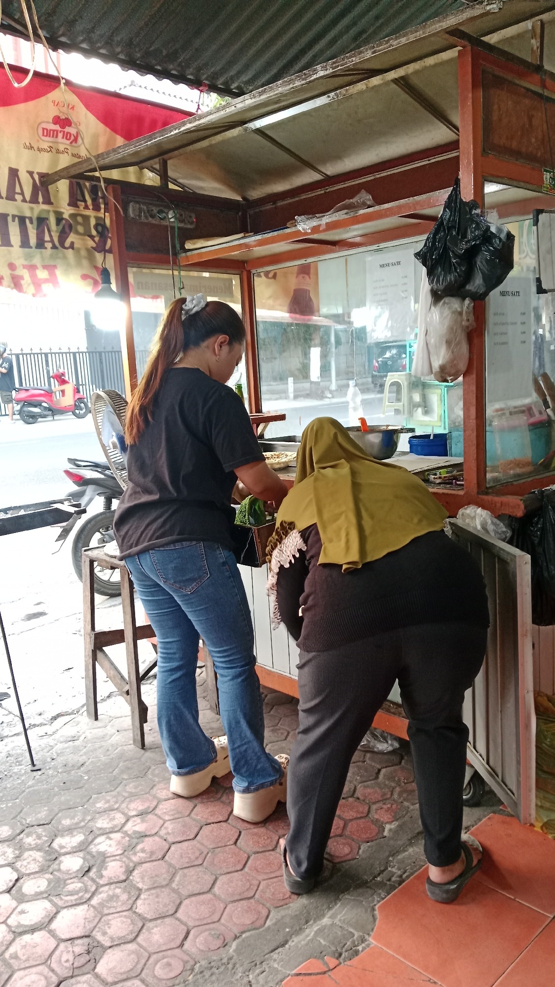 Two women are standing behind a food stall in Blitar, Indonesia. The woman on the left is wearing a black t-shirt, blue jeans, and platform shoes. She has her hair pulled back in a ponytail. The woman on the right is wearing a dark-colored outfit and a green headscarf. She is bent over, looking at something on the counter. The food stall has a metal frame and is covered with a corrugated metal roof. There is a menu visible on the right side of the stall. In the background, a red scooter is parked on the street. The ground is paved with hexagonal tiles.