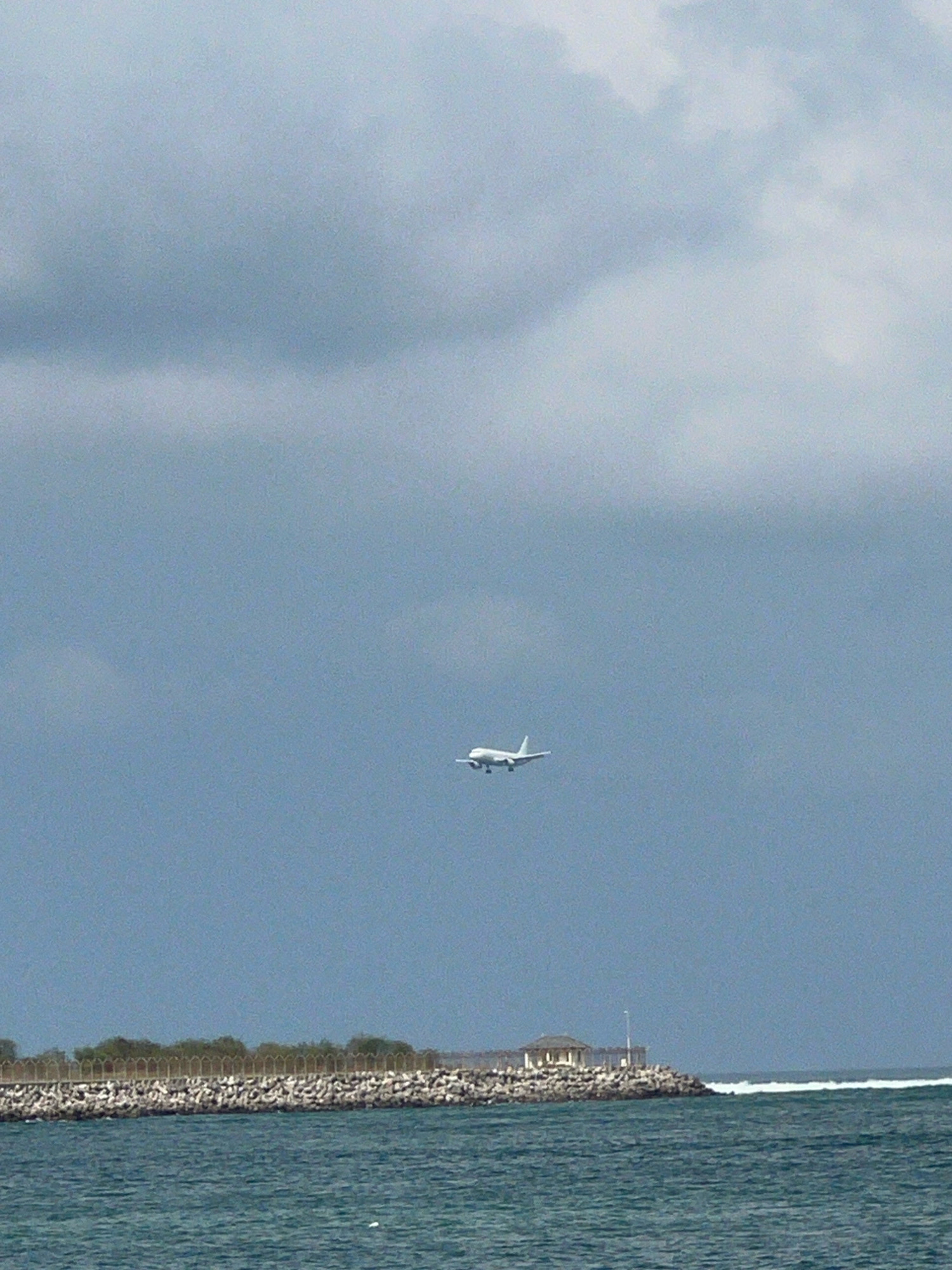 An airplane is visible in the sky above a stone pier and the ocean. The pier is lined with vegetation and a fence, and a small gazebo with a lamppost is situated on its edge. The ocean has moderate waves. The sky is overcast. This scene is in Kuta, Indonesia.
