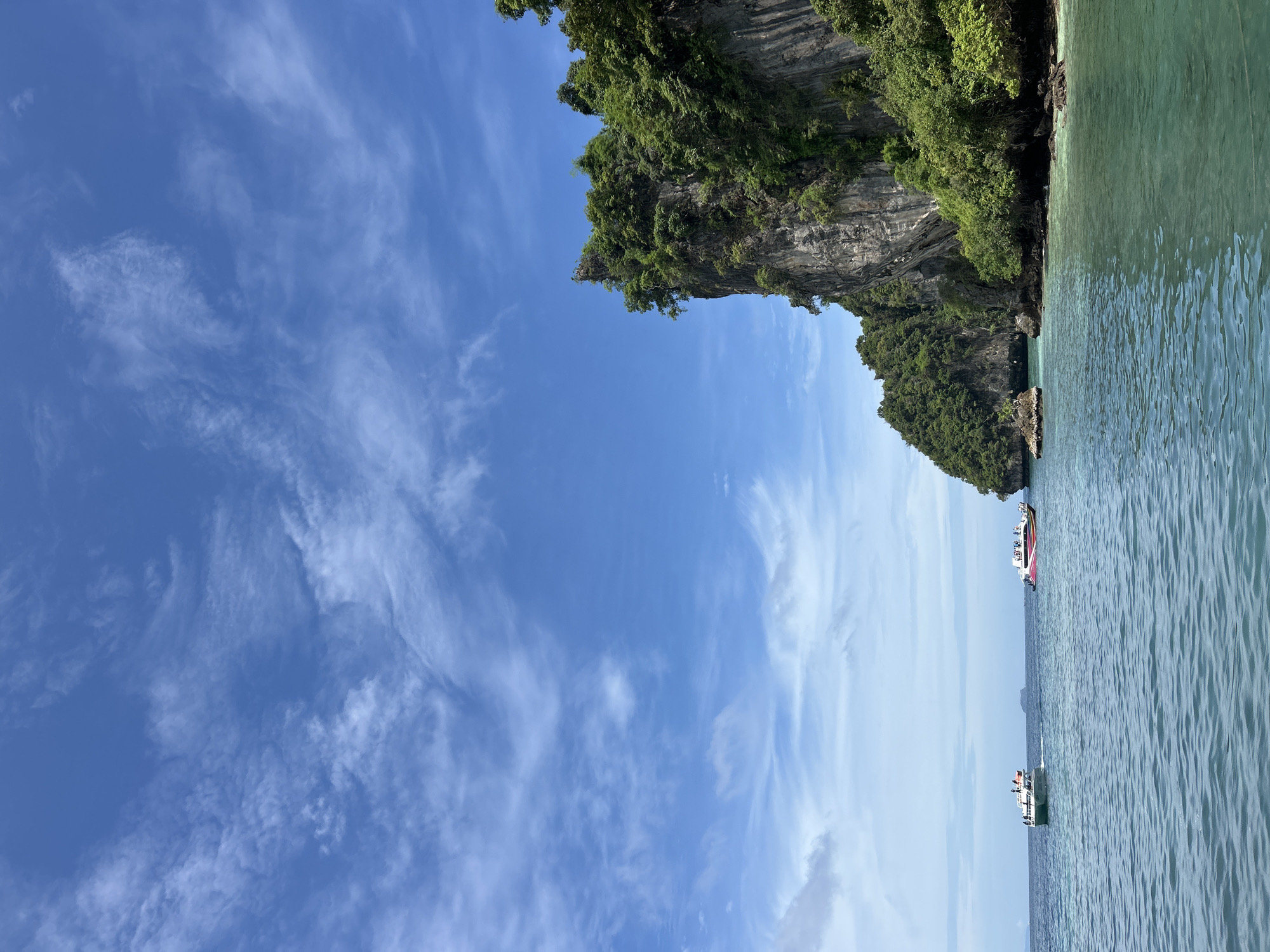 The image depicts a scenic view of islands and the ocean in Ko, Thailand. In the foreground, the clear turquoise water shows gentle ripples. In the middle ground, two boats are visible on the water. The boat on the left is green and white with several people on deck. The boat on the right is white with a colorful striped hull, also carrying passengers. In the background, a large, rocky island covered in lush green vegetation rises dramatically from the sea. The sky above is a vibrant blue, dotted with wispy white clouds, suggesting a clear, sunny day.