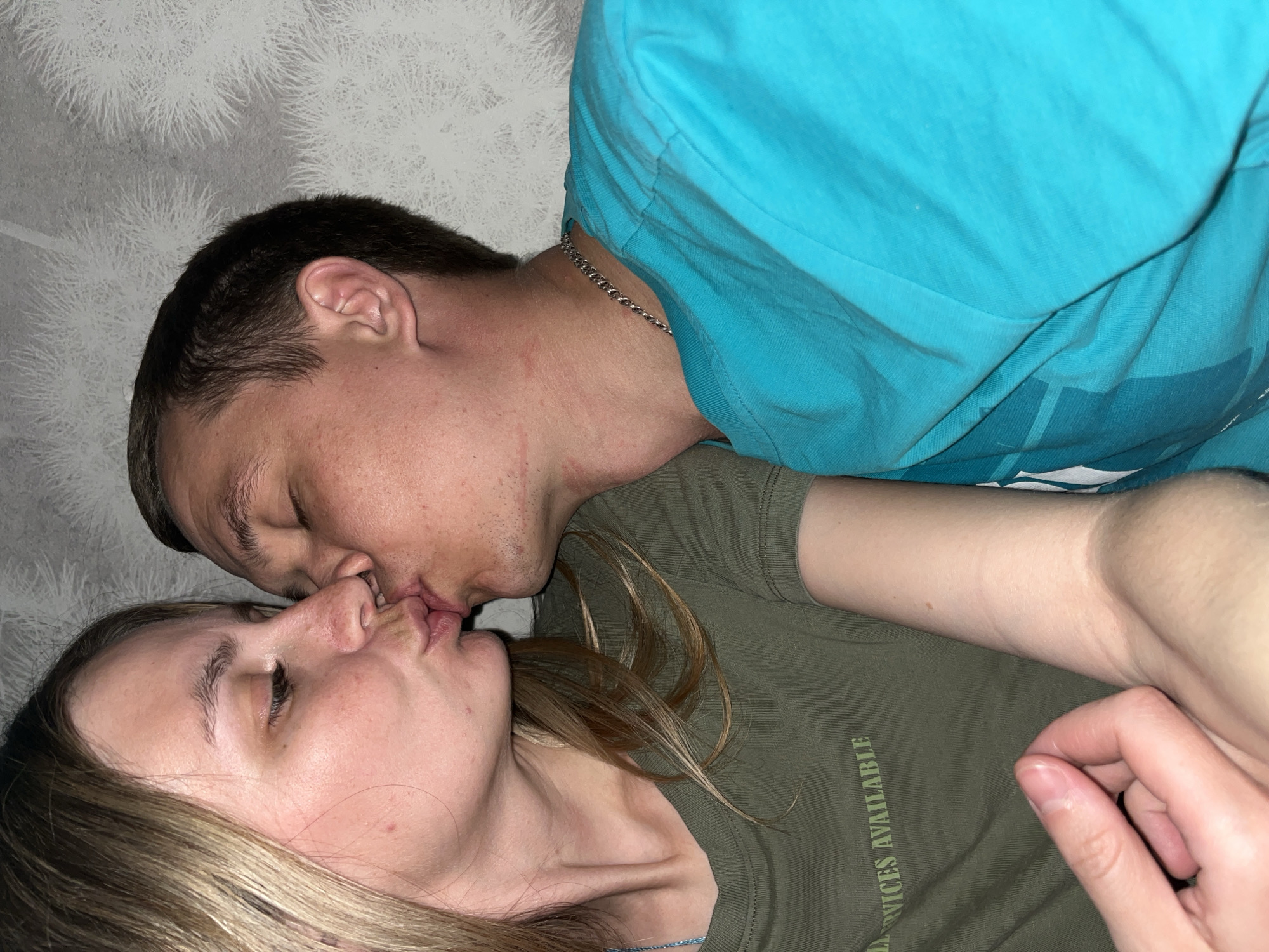 A man and a woman are kissing. The man is wearing a blue t-shirt and a silver chain. The woman is wearing a green t-shirt. The background is a gray and white patterned wall. There is not enough information to determine if the location is Chaikovsky, Russia.