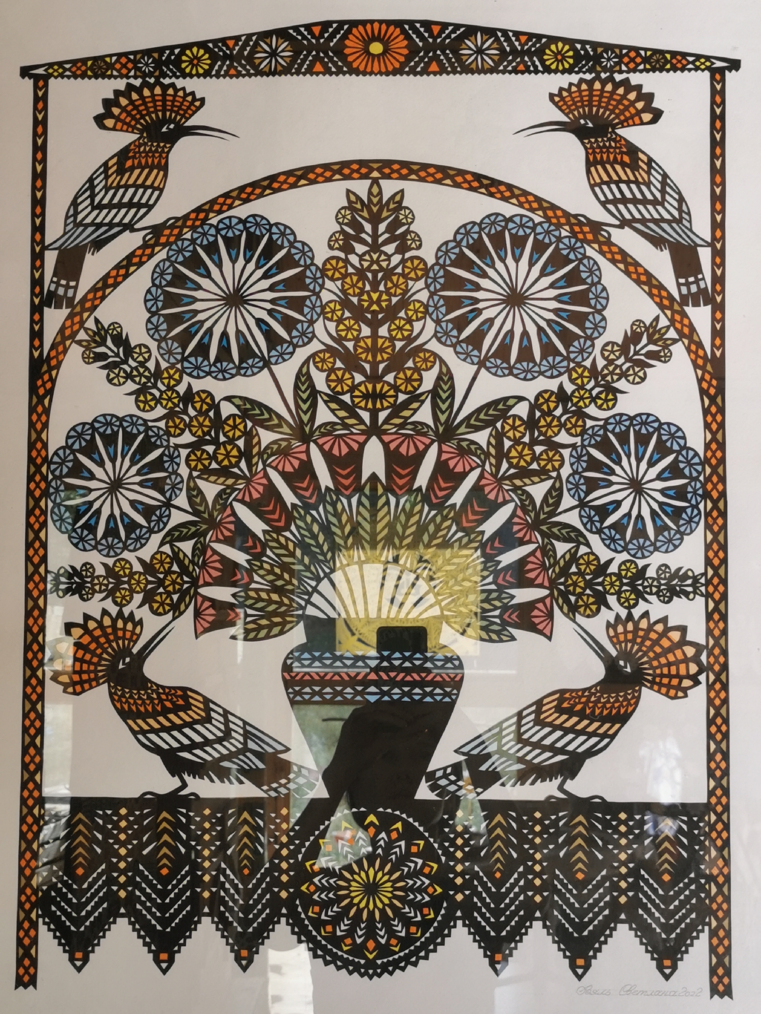 The image is a brightly colored folk art illustration, likely a paper cut-out, depicting stylized hoopo birds perched among floral motifs within a decorative archway. Four hoopo birds are visible, two at the top corners and two at the bottom. They are characterized by their distinctive crests and patterned bodies rendered in black, white, and orange. The central part of the illustration features a vase from which a stylized tree or bouquet of flowers emerges. This central arrangement incorporates circular, star-like floral elements in blue and white, as well as smaller yellow blossoms. The entire composition is framed by decorative borders with geometric patterns in black, orange, and yellow. A signature and date, "Barys Cemjancov 2022," is present in the bottom right corner.
This artwork is located in the village of Ščarčoŭski sielski Savet, Belarus. The image is taken from a slightly upward angle, suggesting it is hanging on a wall or displayed upright. The lighting indicates a well-lit indoor environment, with subtle reflections on the surface of the artwork, possibly from glass covering. The background of the artwork is white.